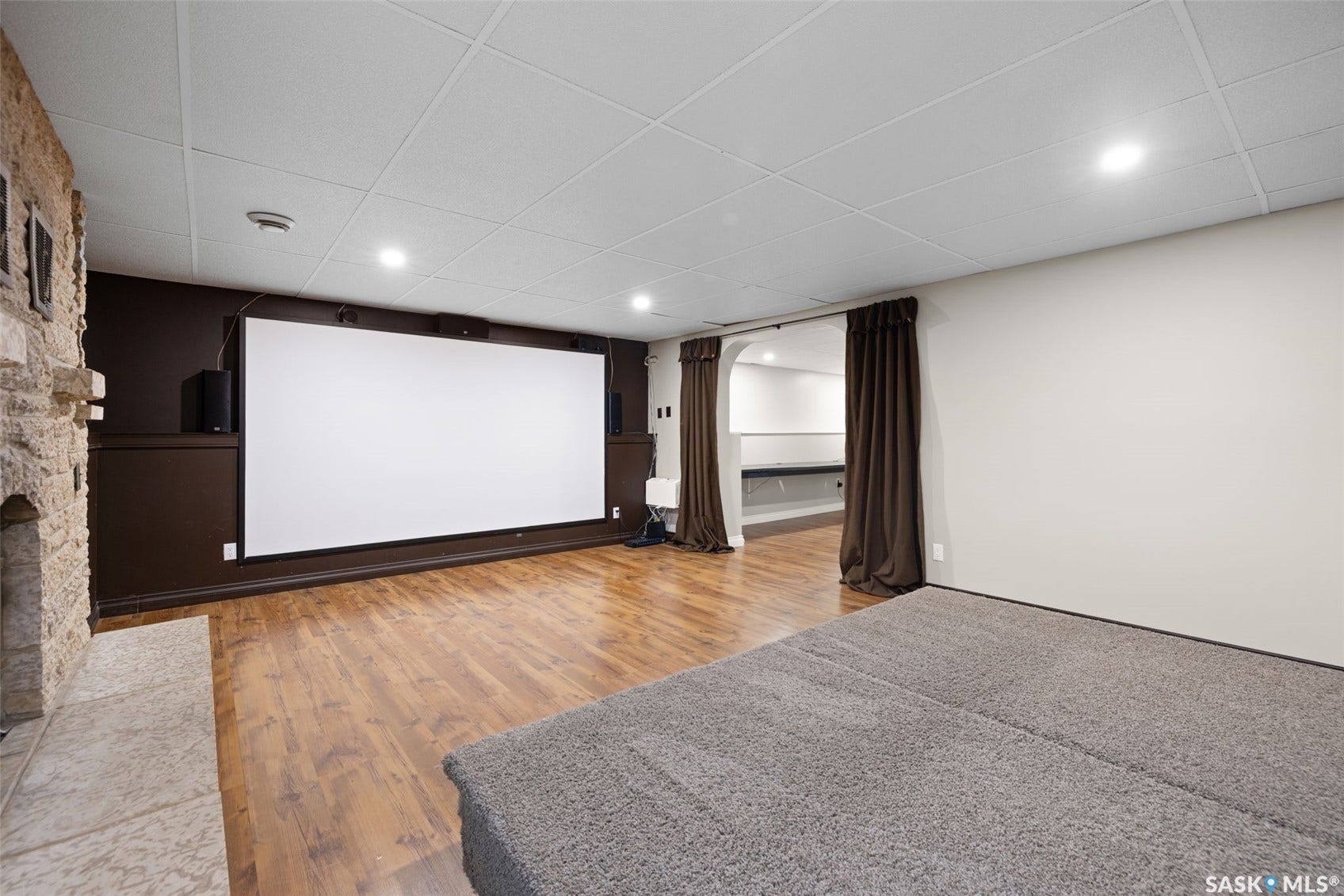 Spacious entertainment area featuring a large projection screen, stone-clad fireplace with hearth, wood-finish flooring, recessed lighting, and a suspended ceiling