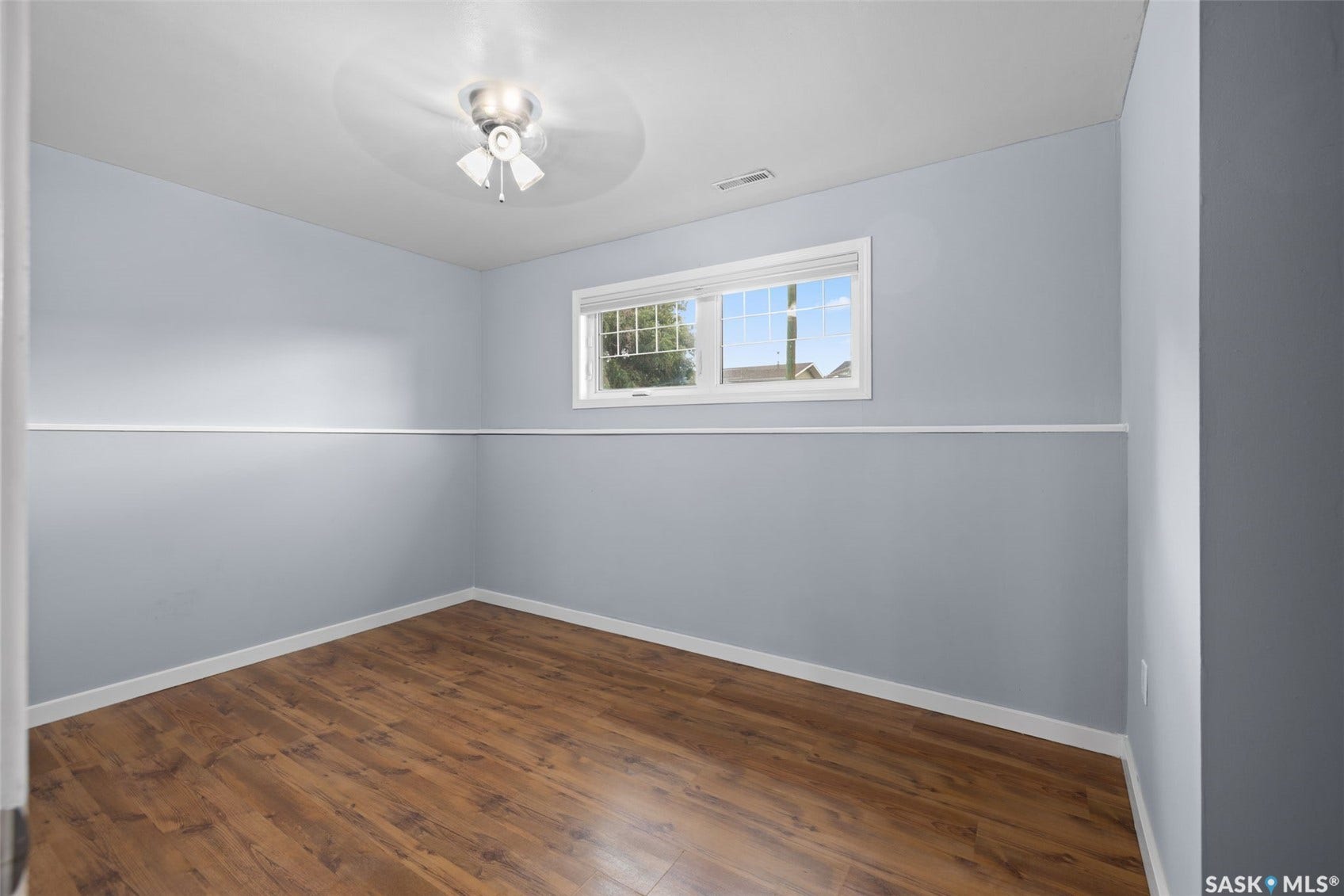 Wood-finish flooring, light blue walls, white trim, window with blinds, and a ceiling fan with integrated lighting