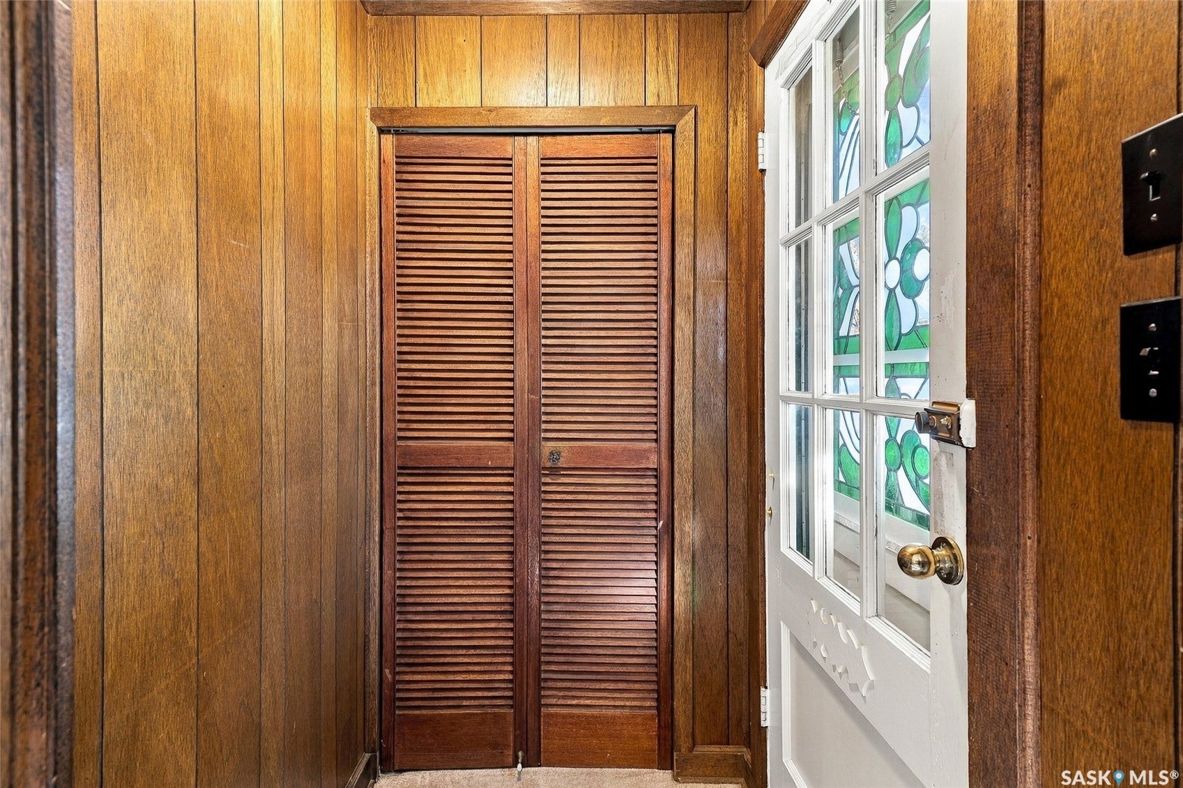 Elegant wood-paneled hallway with louvered closet doors, complemented by a white-trimmed exterior door with decorative glass panels and brass hardware