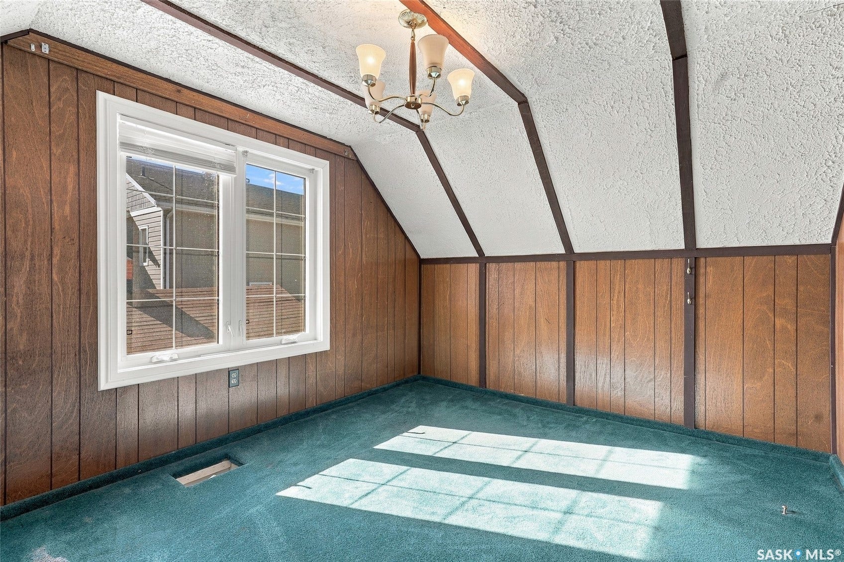 Wood paneling, vaulted ceiling with exposed beams, carpet flooring, chandelier lighting, and a multi-pane window