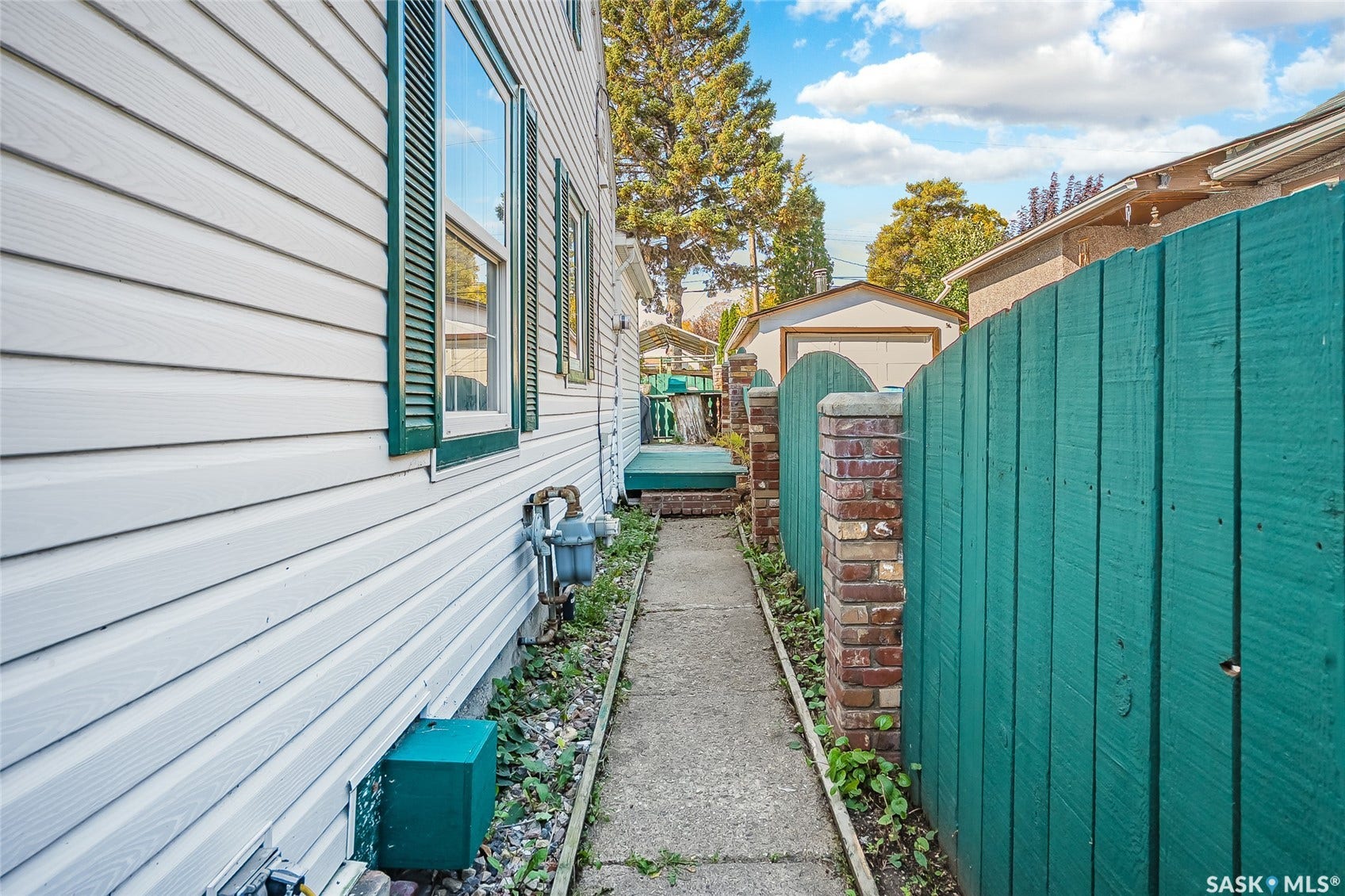 White horizontal siding with green window shutters, concrete walkway, brick pillars, and a green privacy fence