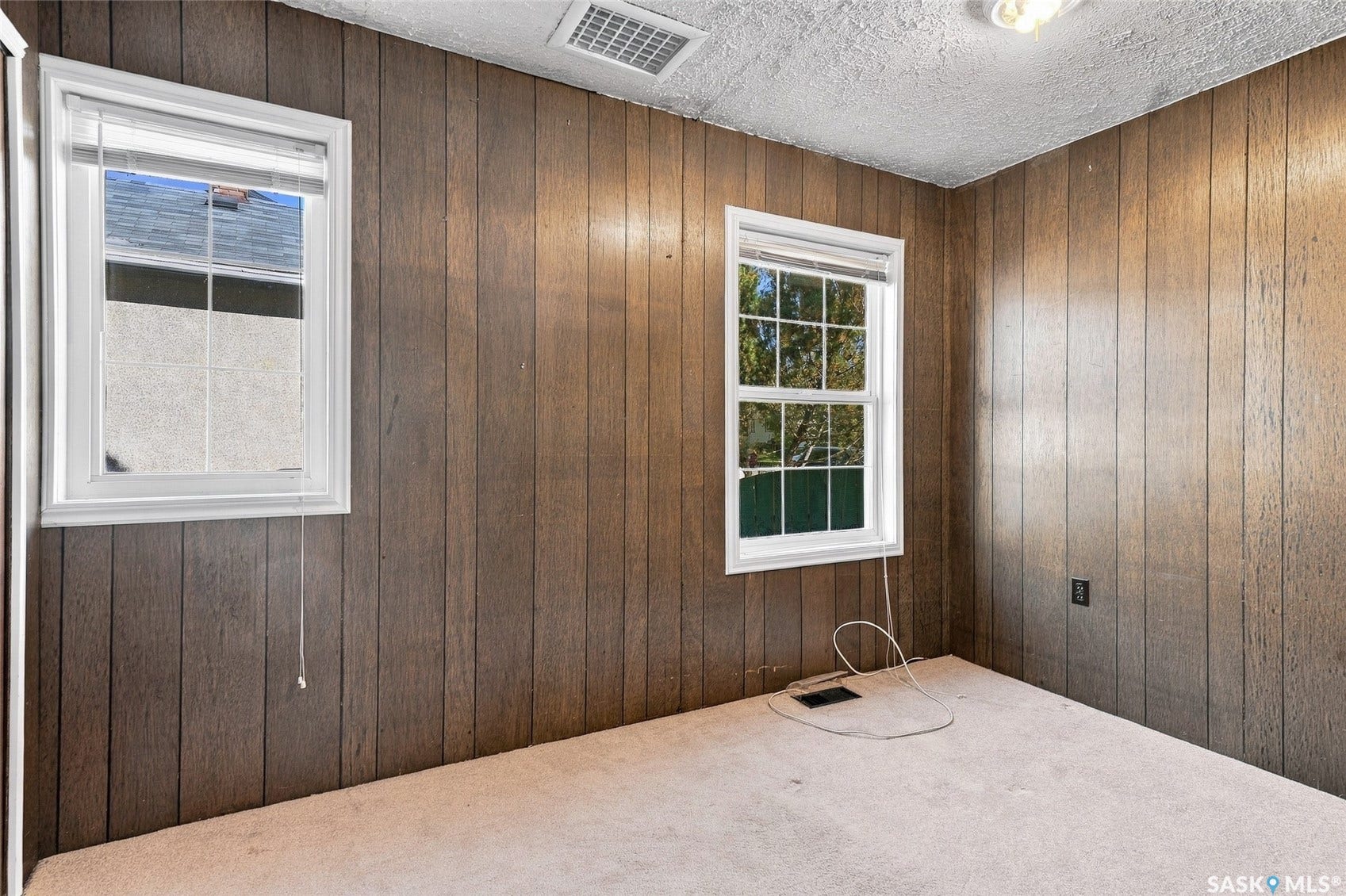 Brown wood-paneled walls, two white-framed windows, textured ceiling, and light-colored carpeting