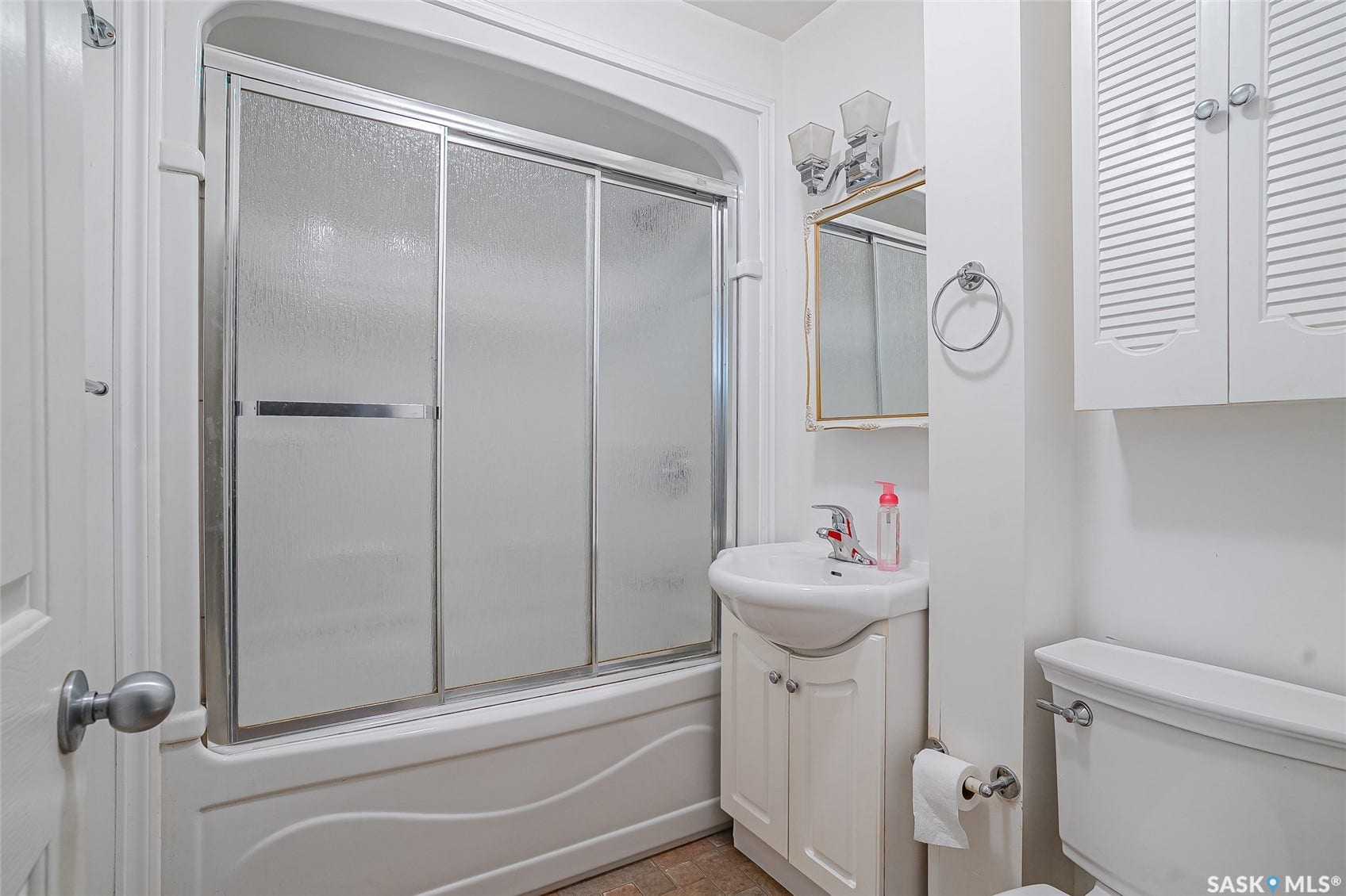 Bathtub with sliding frosted glass enclosure, white pedestal sink with cabinet storage, wall-mounted vanity mirror, and overhead cabinet with louvered doors