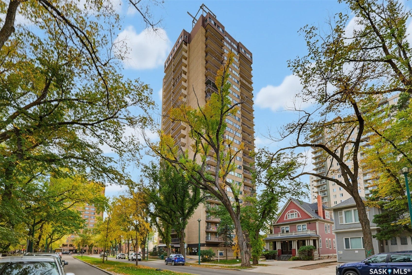 Tall residential building with balconies, surrounded by mature trees and residential structures