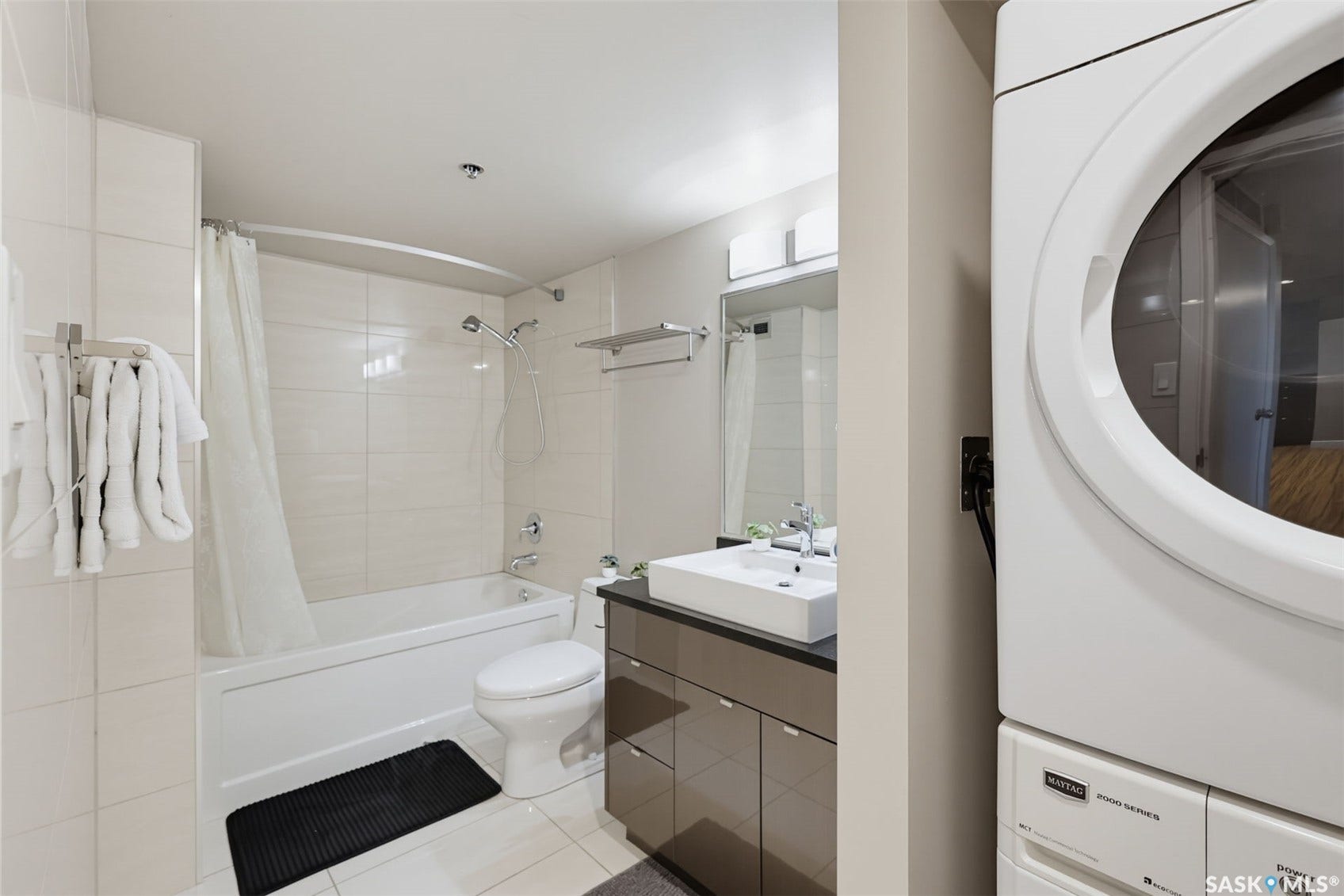 Stacked laundry appliances, white rectangular vessel sink, dark countertop, vanity with wood-finish cabinetry, and a full bathtub with shower