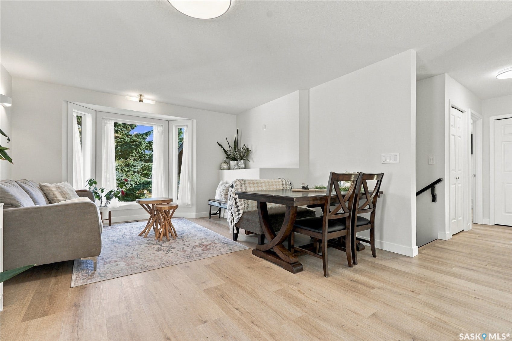 Spacious living area featuring light wood-finish flooring, a bay window with a garden backdrop, and white walls