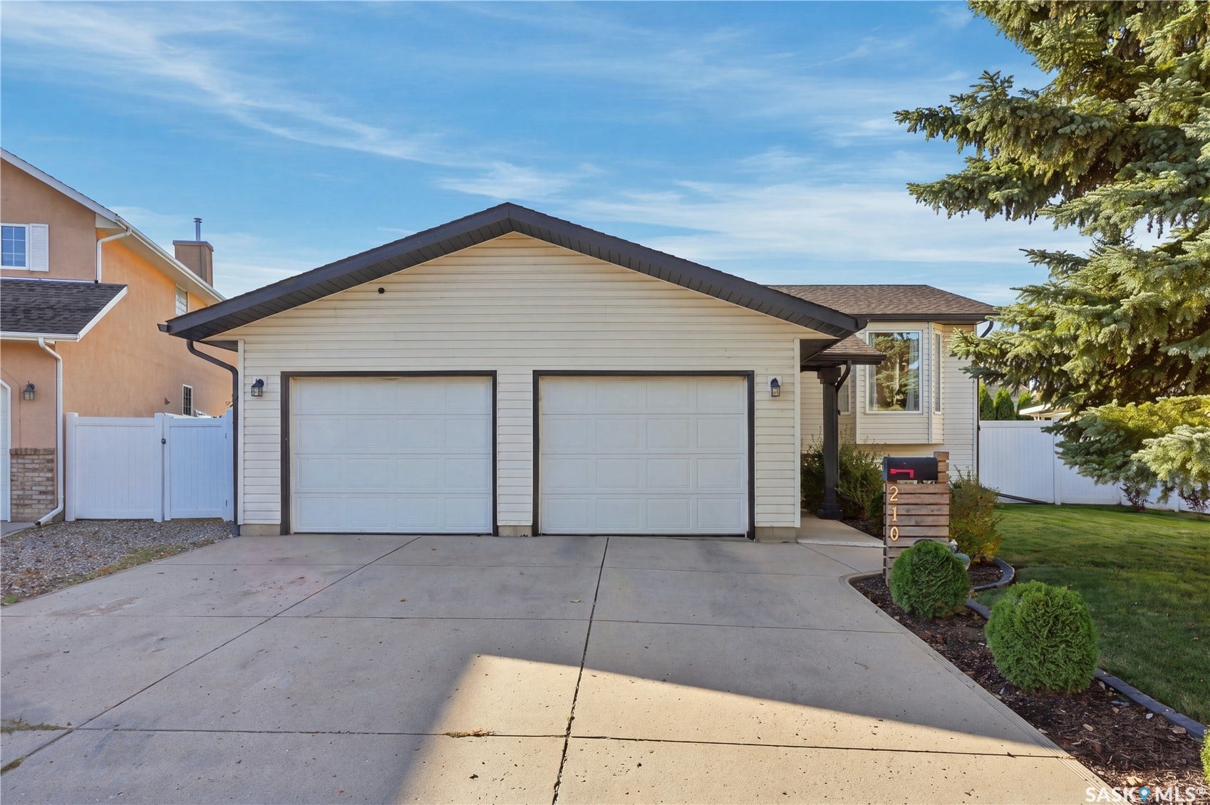 Two-car garage with white doors, light-colored siding, dark trim, concrete driveway, and mature landscaping