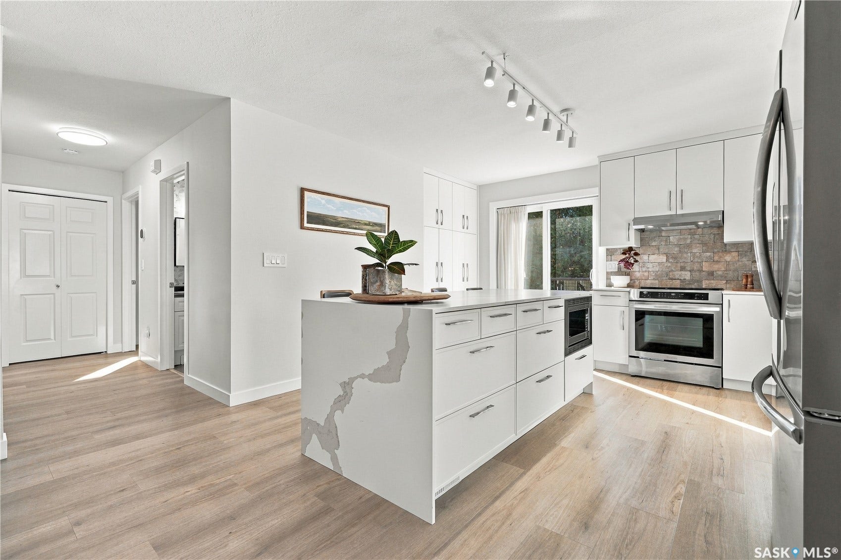 Modern kitchen featuring an island with a waterfall countertop, white cabinetry with silver hardware, stainless steel appliances, and wood-finish flooring
