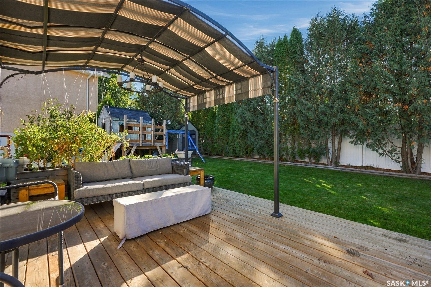 Expansive wood-finish deck with a striped canopy, overlooking a manicured lawn and mature privacy trees