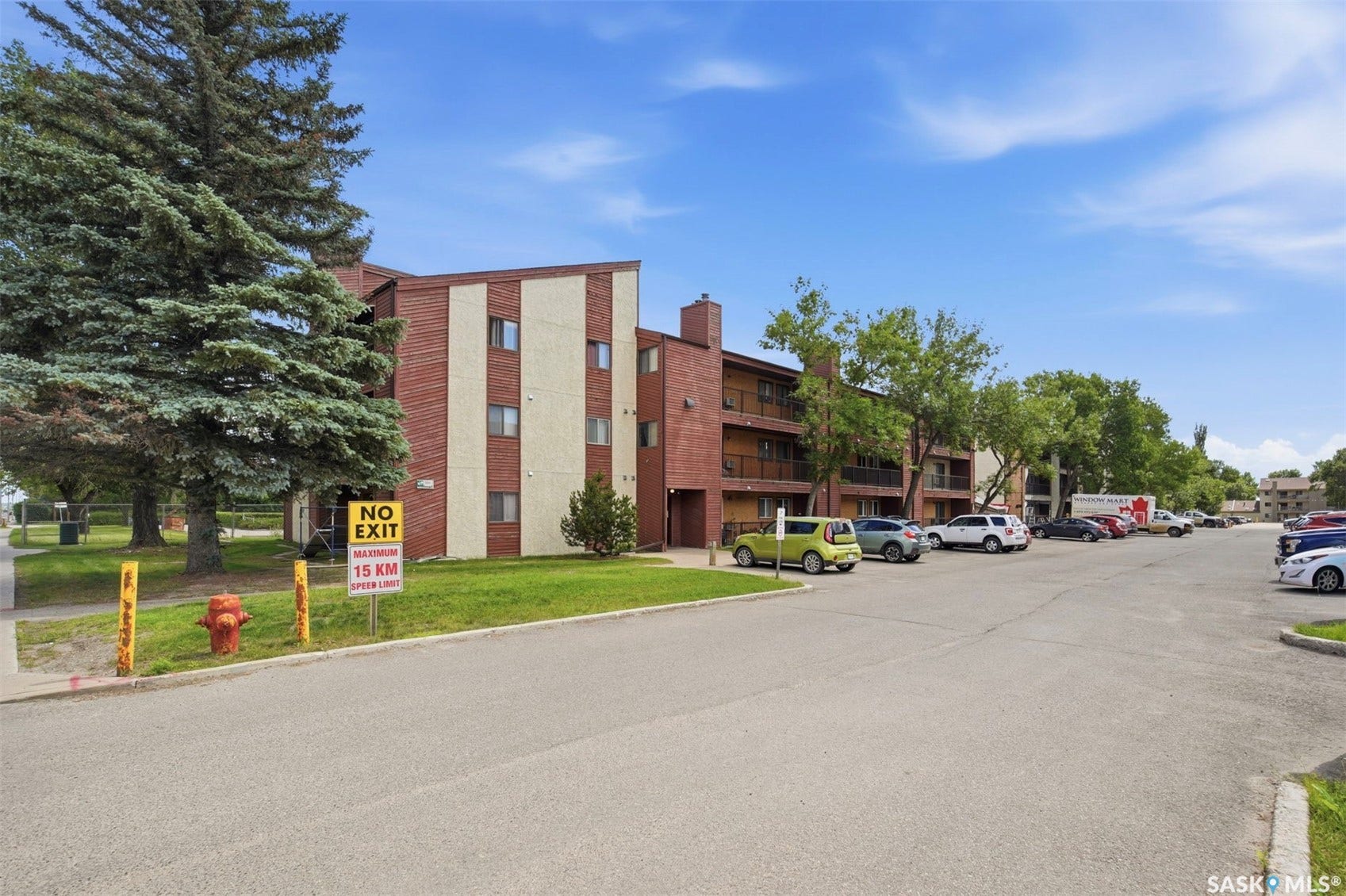Brick exterior with balconies, surrounded by mature trees, and an asphalt parking lot