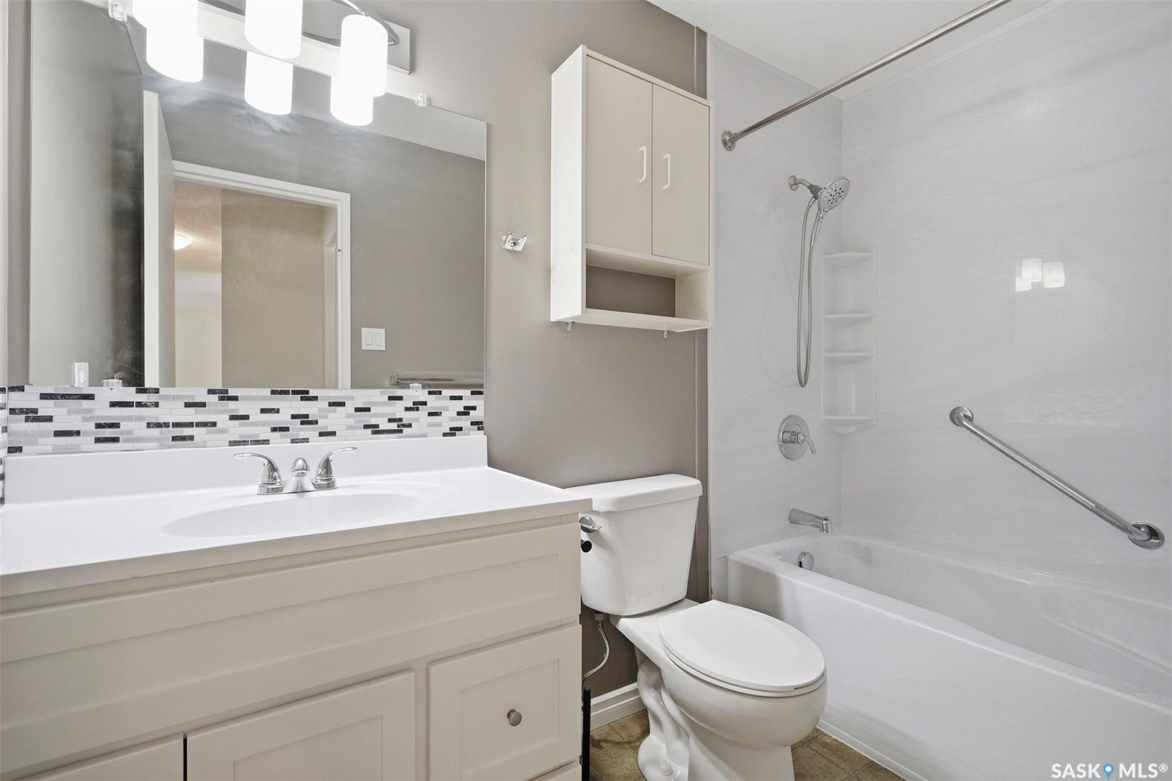 White vanity with an integrated sink, mosaic tile backsplash, built-in wall cabinet, and a white bathtub with grab bars