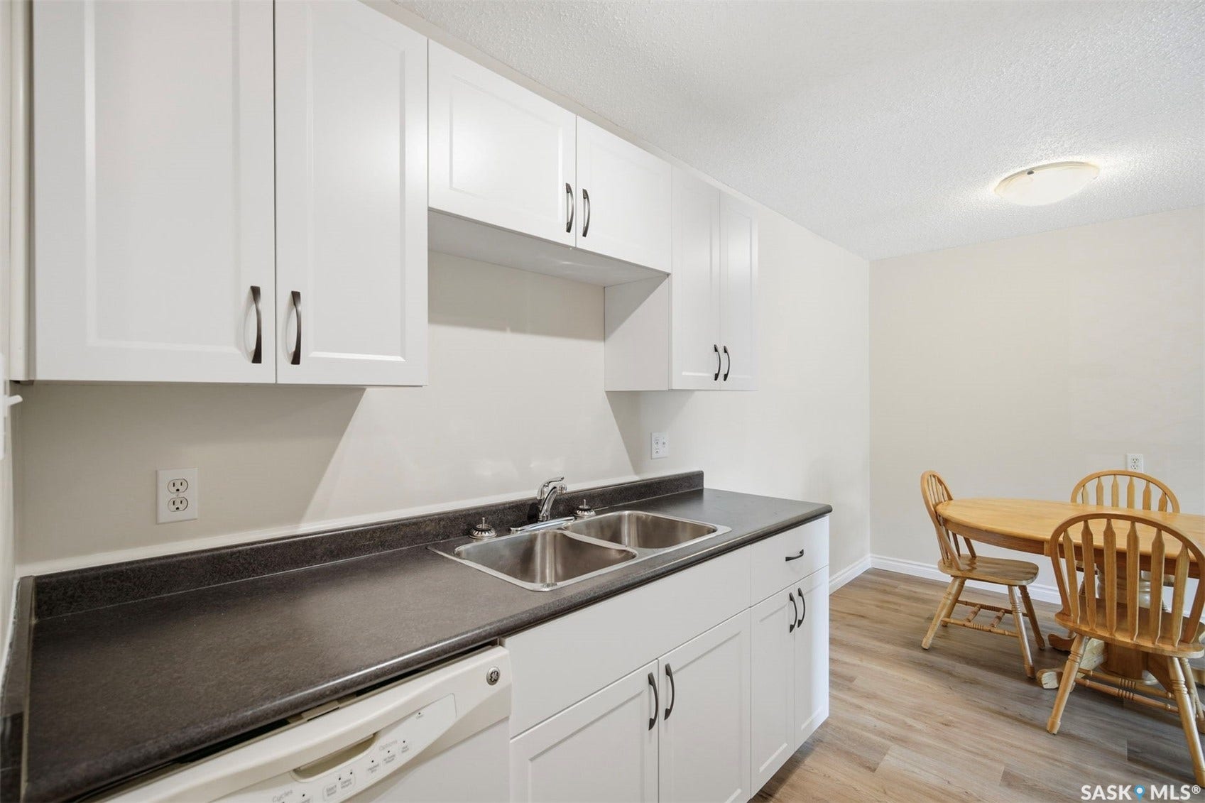 White shaker-style cabinetry, dark countertops, a double basin stainless steel sink, wood-finish flooring, and a flush mount ceiling light fixture
