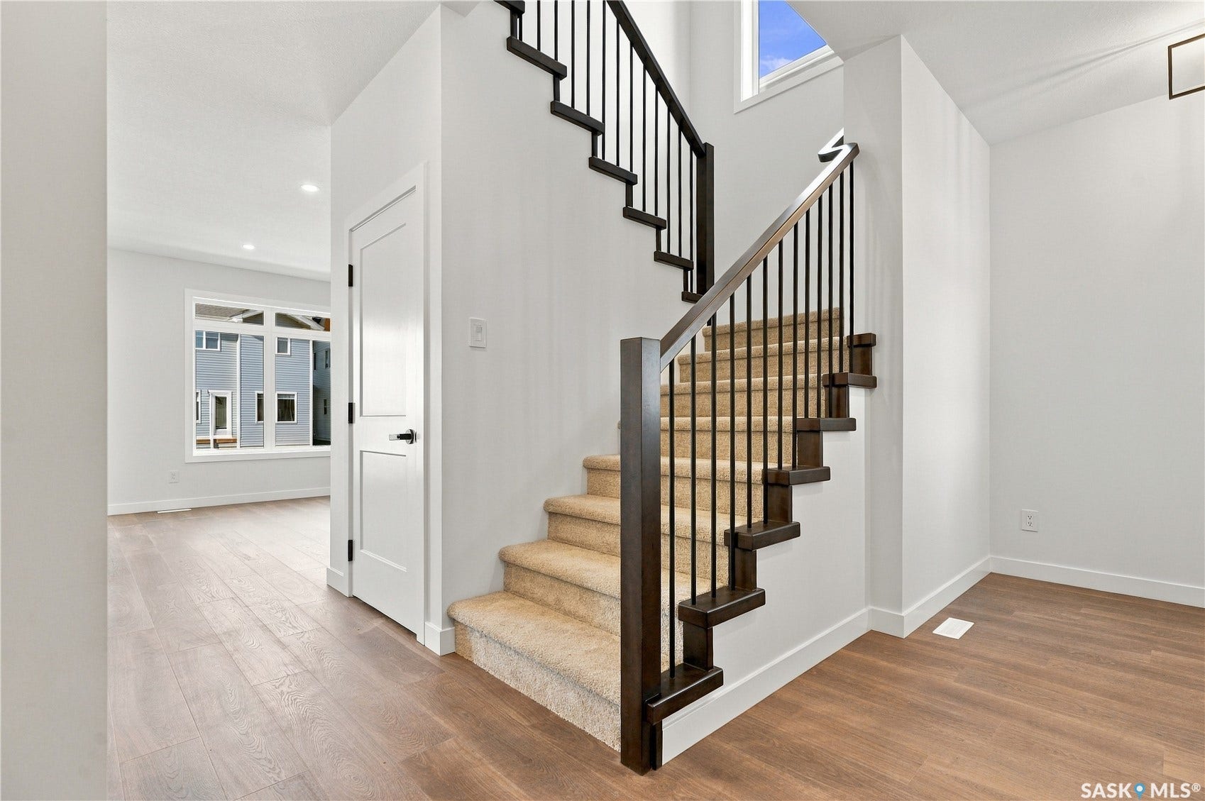 Wide-plank wood-finish flooring, staircase with dark wood newel posts and metal spindles, carpeted stair treads, white interior doors with dark hardware, and a large multi-pane window