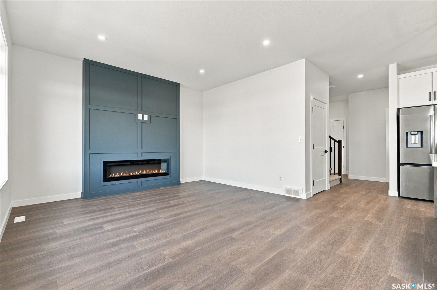 Expansive living space featuring wood-finish flooring, a built-in electric fireplace with a teal accent surround, recessed lighting, and a stainless steel refrigerator