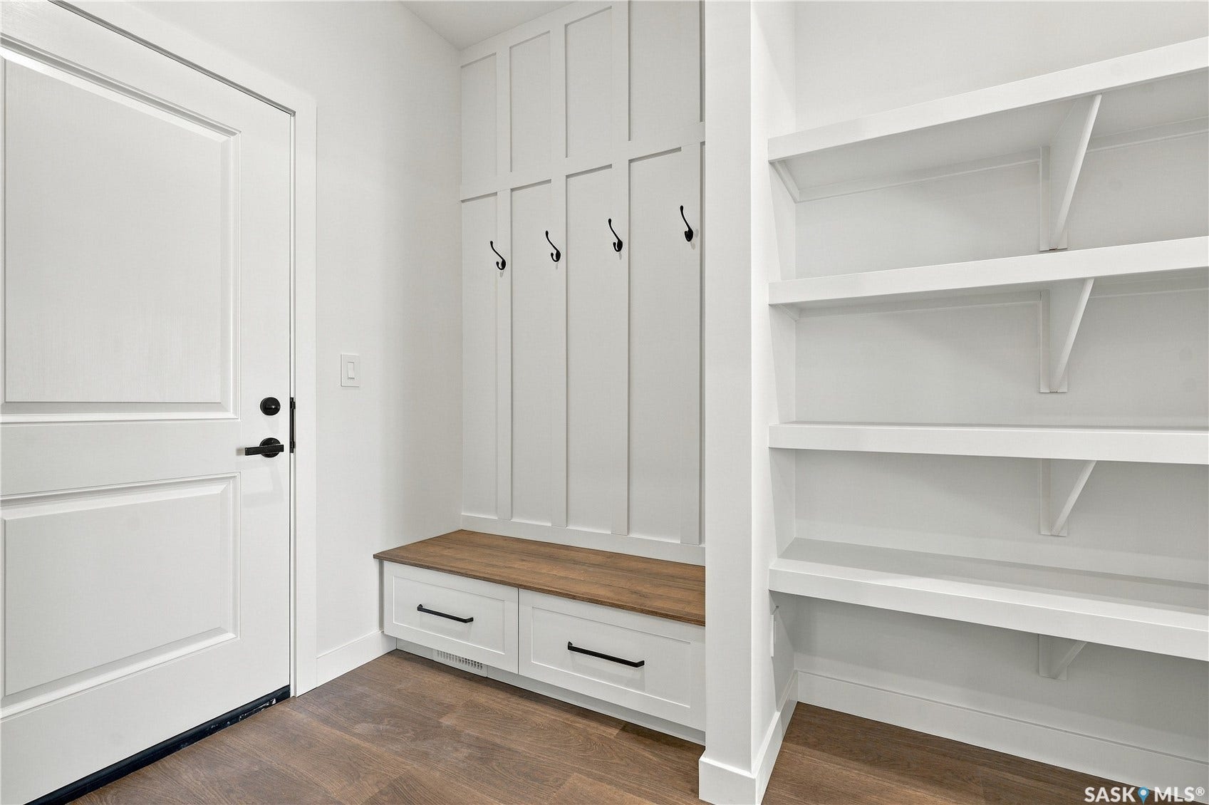 Entryway featuring wood-finish flooring, white paneled door with black hardware, built-in bench with drawers, wall-mounted coat hooks, and open shelving