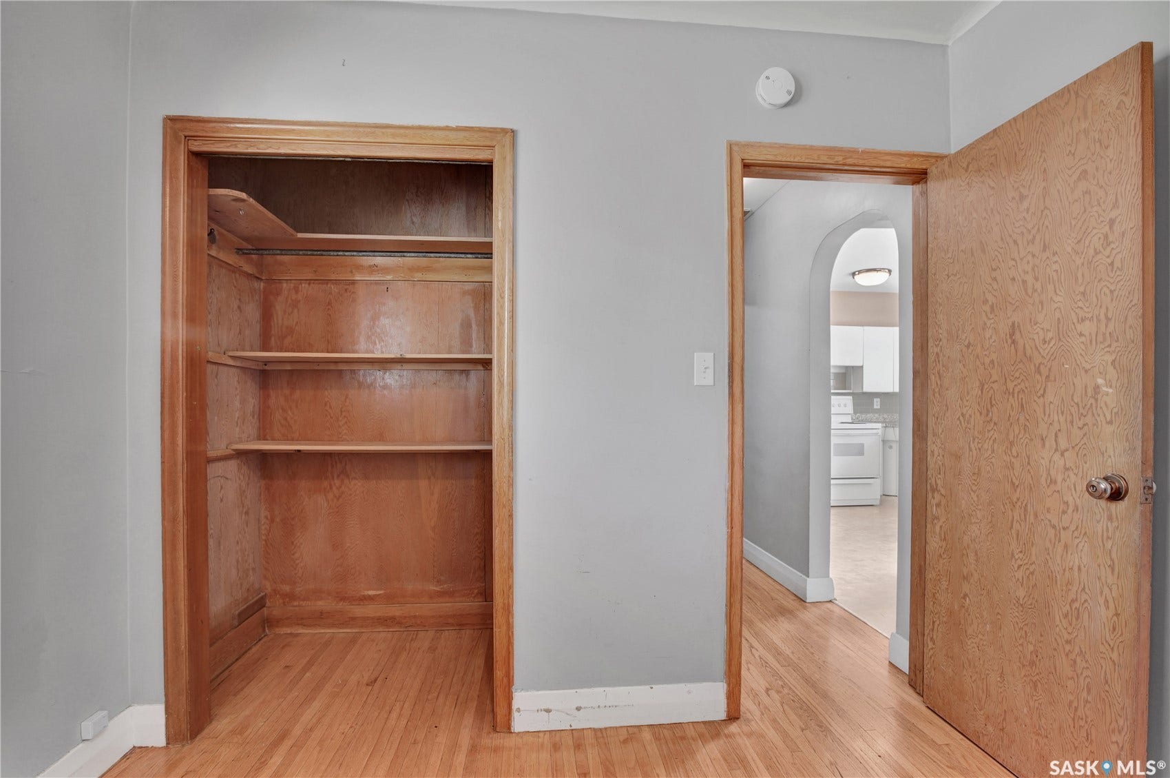 Hardwood flooring, built-in shelving, wood-paneled closet interior, wood door with round knob, arched hallway entrance