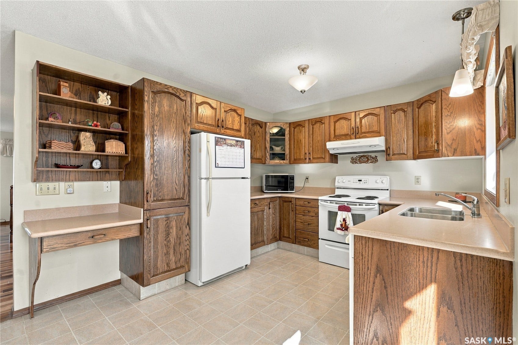 Wood-finish cabinetry with display shelving, white appliances, and a built-in desk area, complemented by light-toned countertops and tile flooring