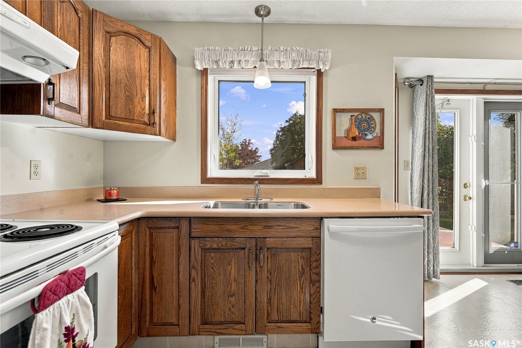Kitchen with wood cabinetry, L-shaped countertop, stainless steel sink, and white appliances