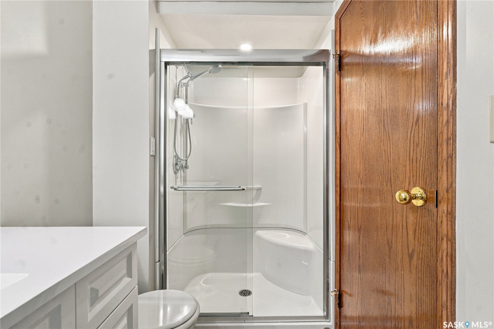 White vanity with integrated sink and chrome faucet, glass-enclosed shower with built-in seating, and wood-paneled door with brass hardware