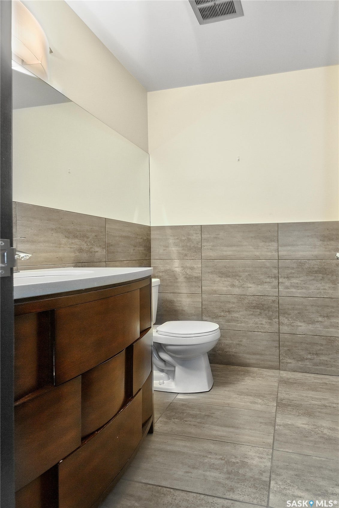 Curved wood-finish vanity with white countertop, full-width mirror, and large format floor and wall tiling