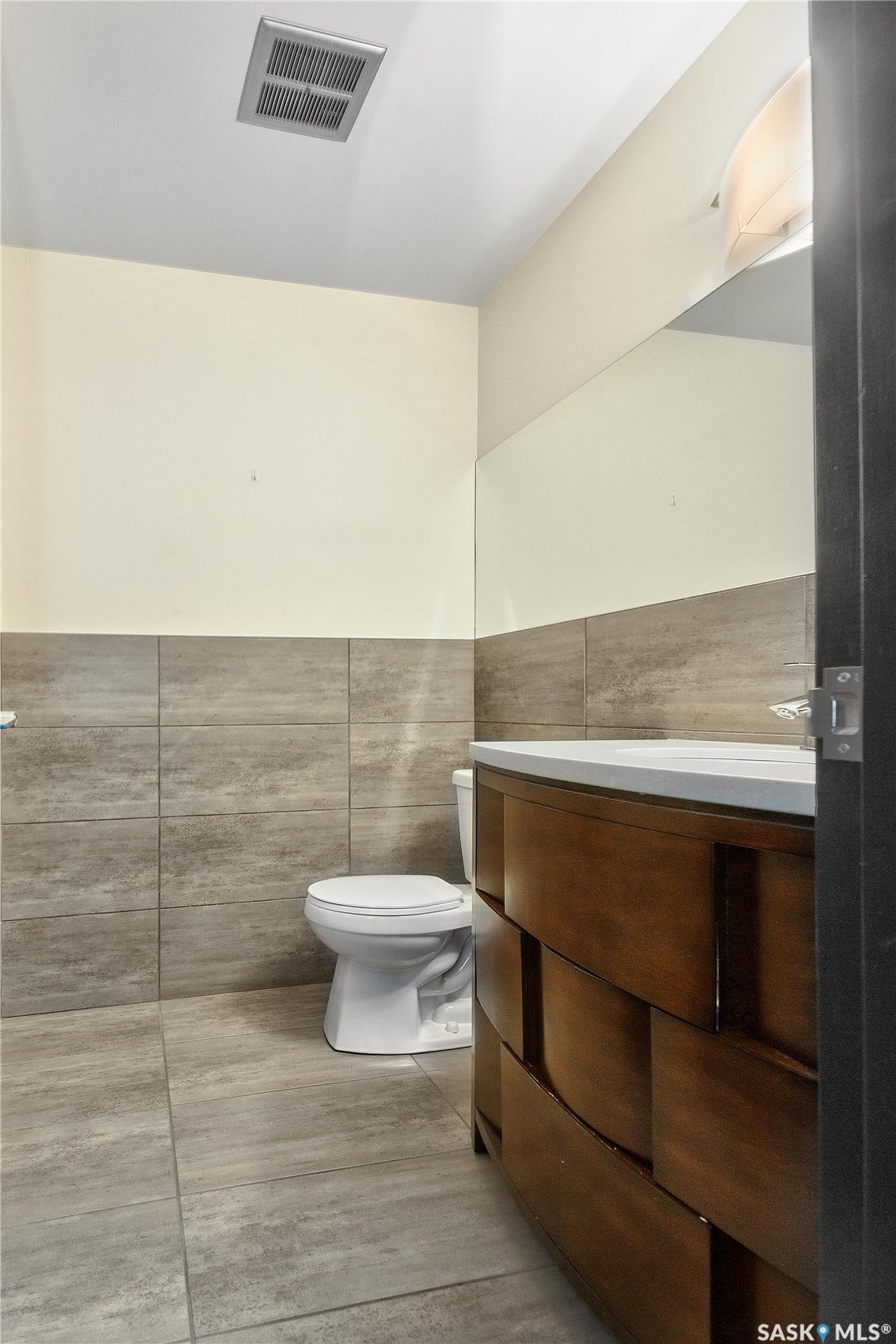 Porcelain tile flooring, tile wainscoting, wood-finish vanity with countertop, wall-mounted mirror, and wall-mounted light fixture