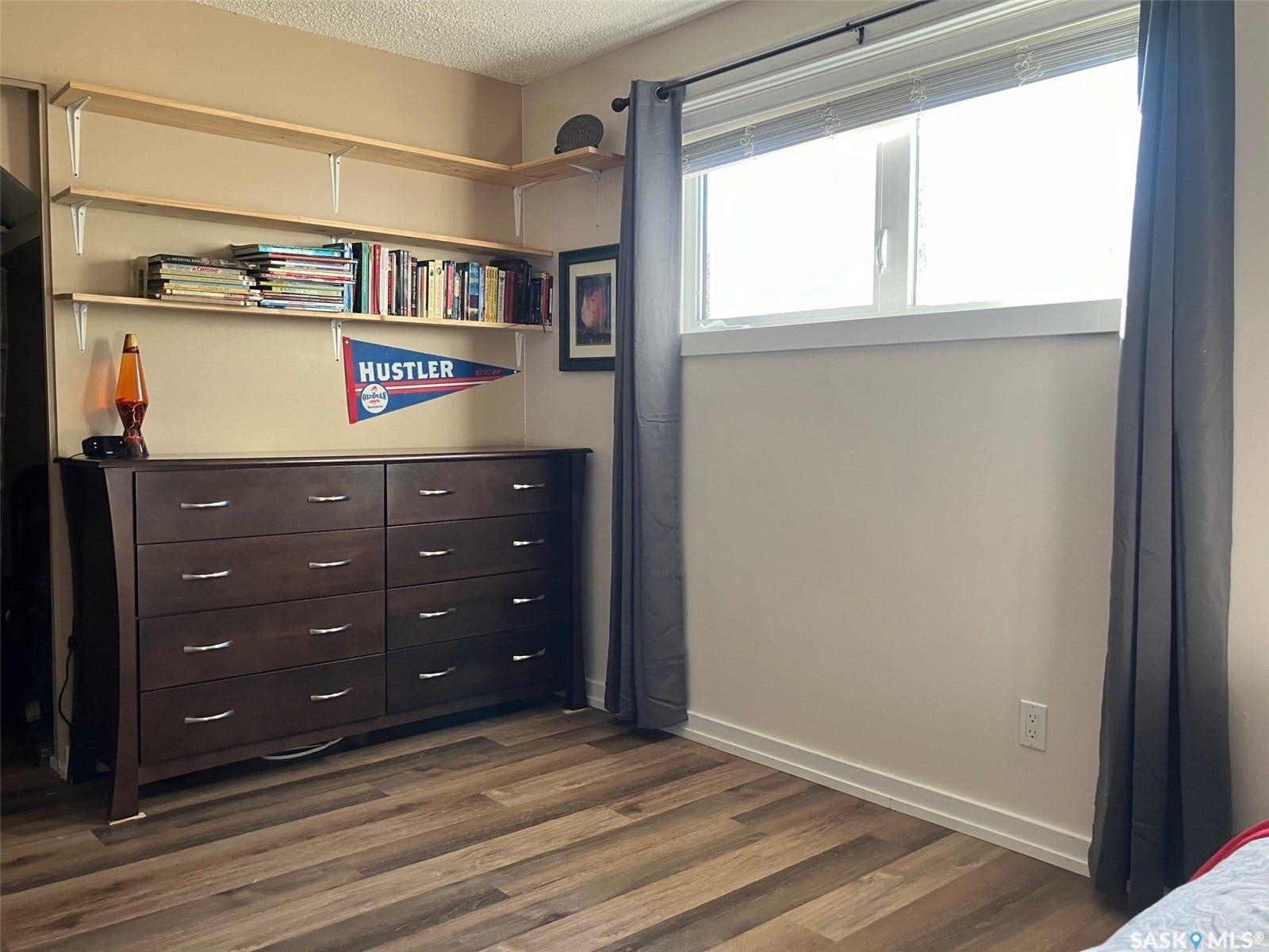 Wood-finish flooring, corner wall-mounted shelving, and a large window with privacy blinds