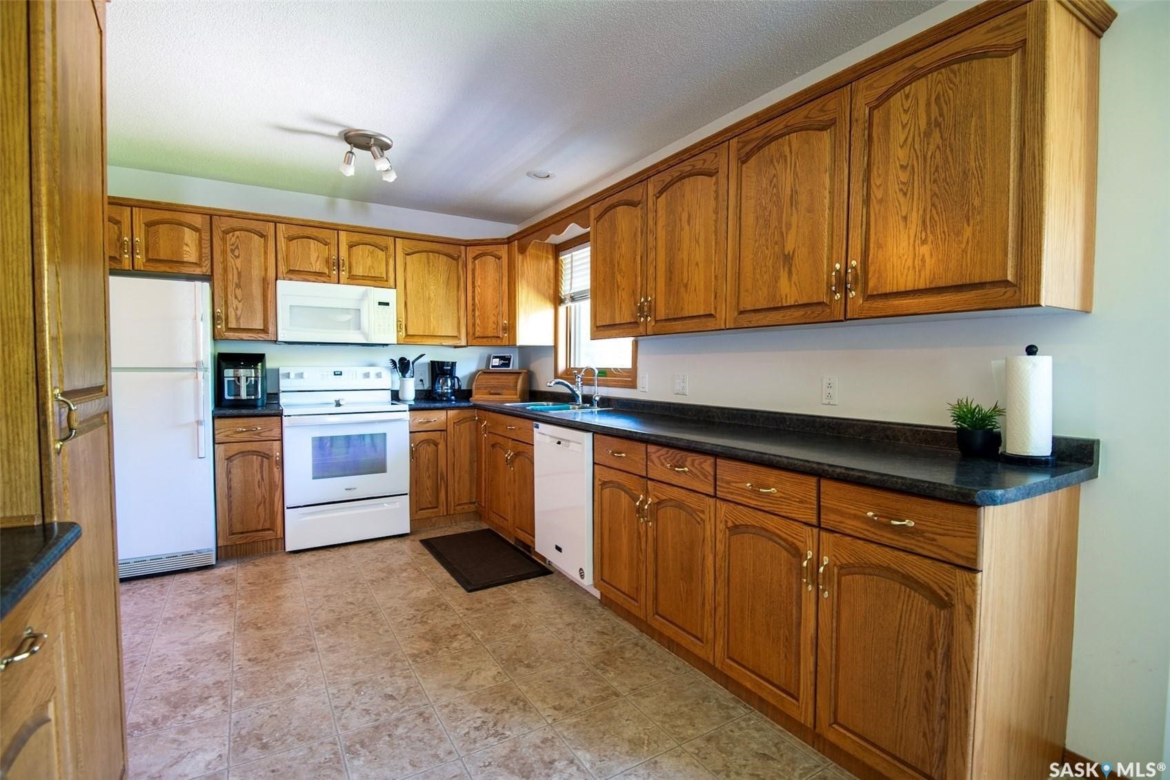 Kitchen featuring wood-finish cabinetry, dark countertops, white appliances, and tiled flooring