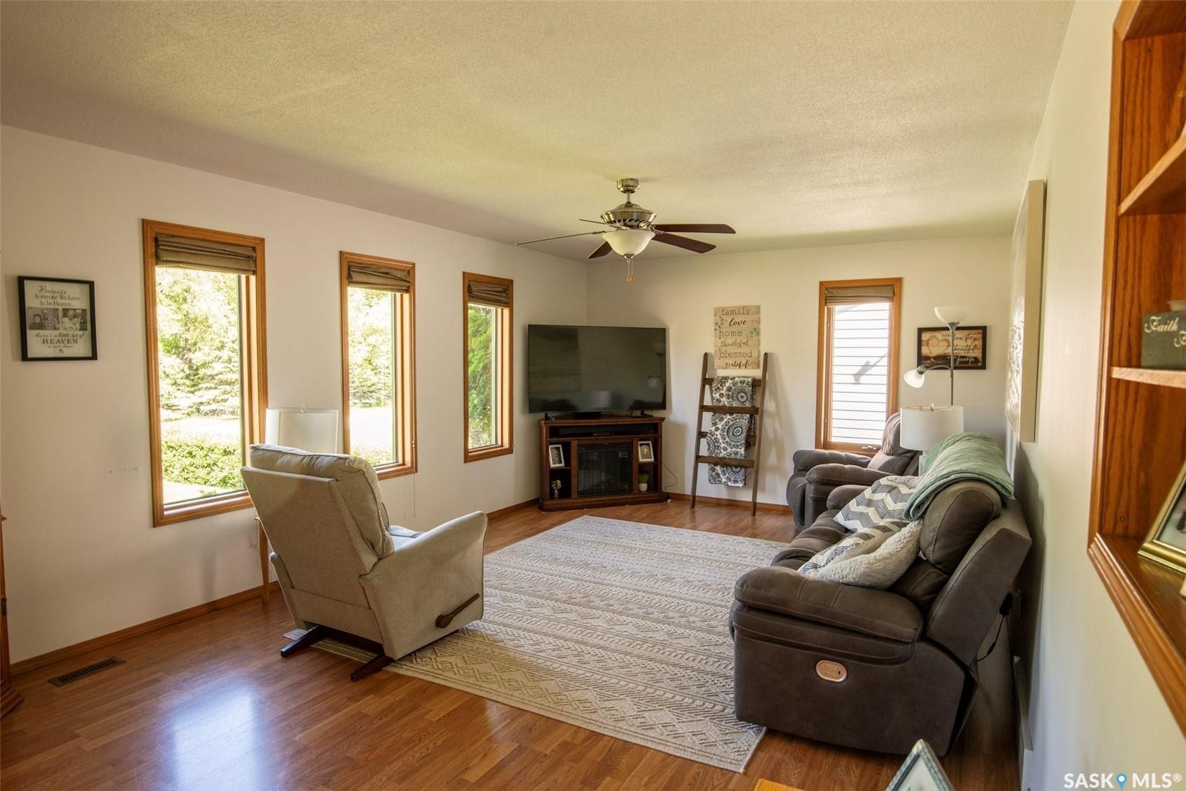 Spacious living area featuring laminate-finish flooring, a ceiling fan, and multiple windows with wood trim