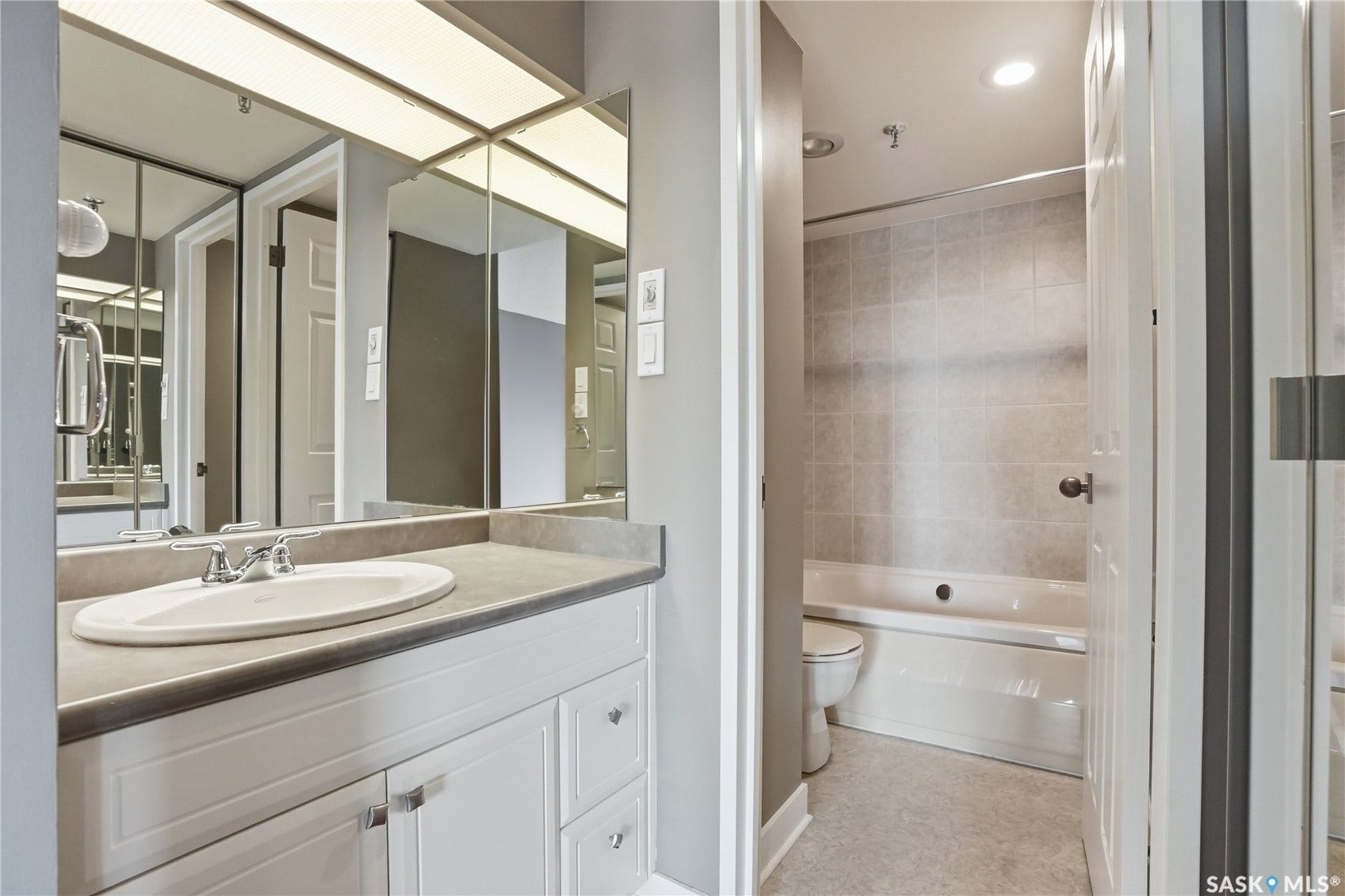 Vanity with white cabinetry and oval sink, integrated countertop, overhead fluorescent lighting, full-wall vanity mirror, and tiled shower/tub combination