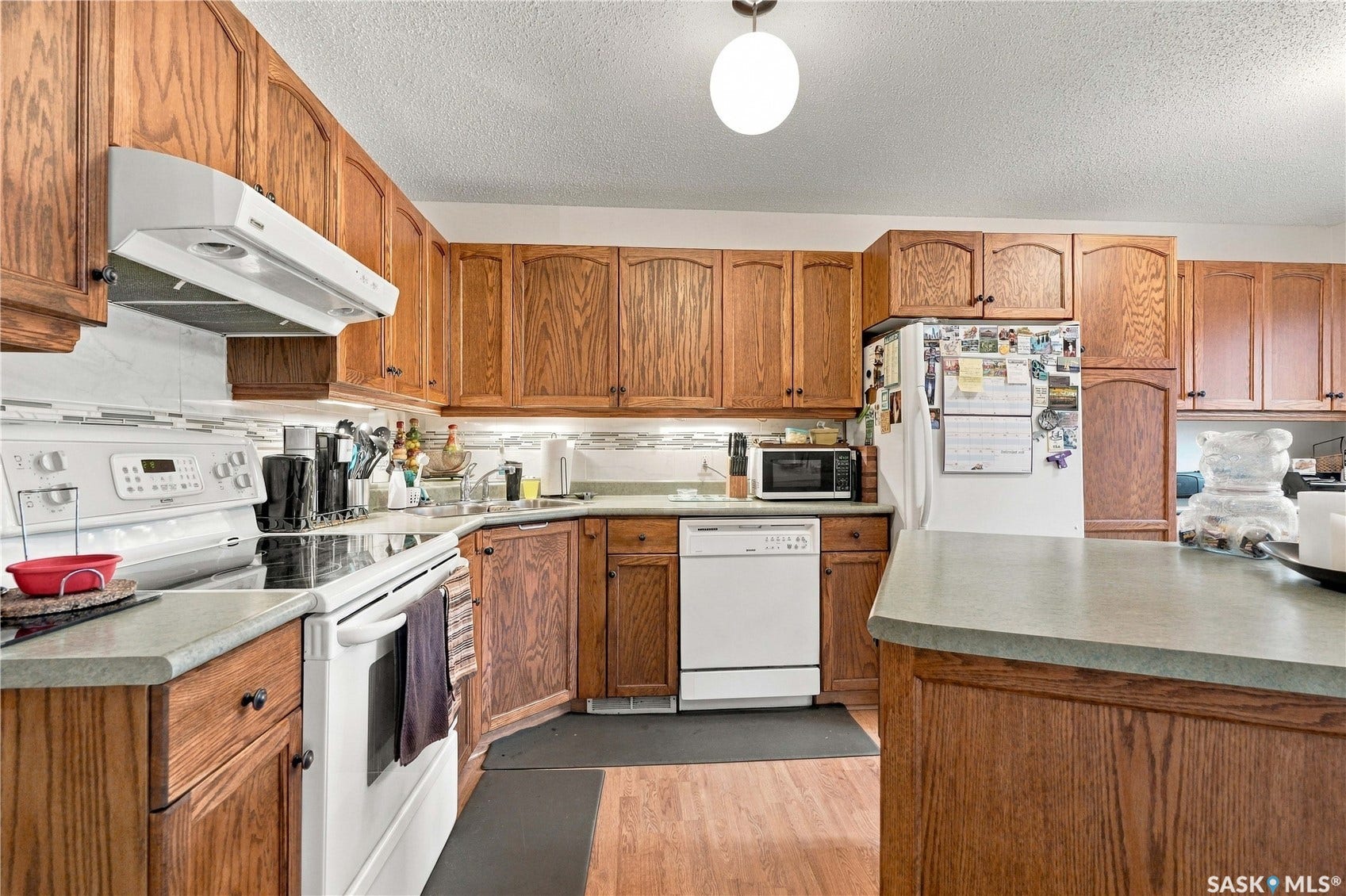 Wood-finish flooring, abundant wood-finish cabinetry, light-toned countertops, white tile backsplash, and white appliances