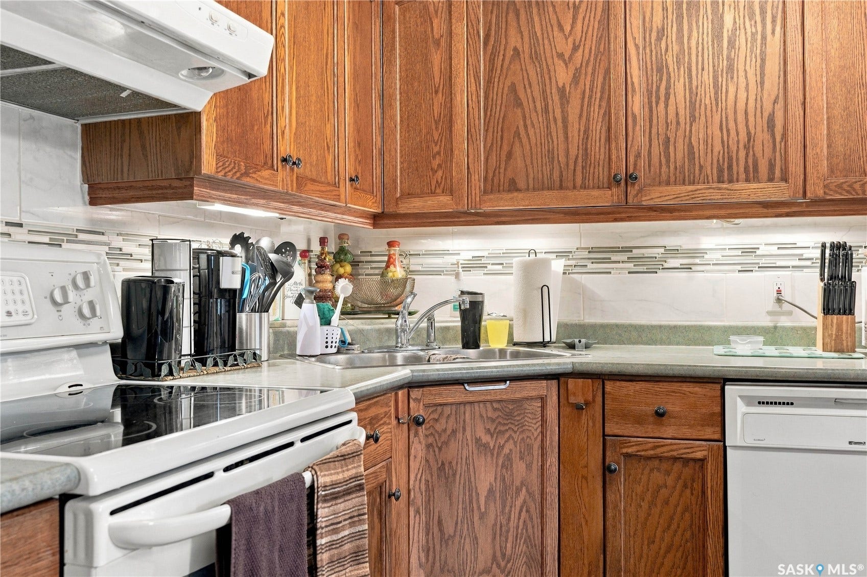 Wood cabinetry, corner sink, white electric range, mosaic tile backsplash, and built-in dishwasher