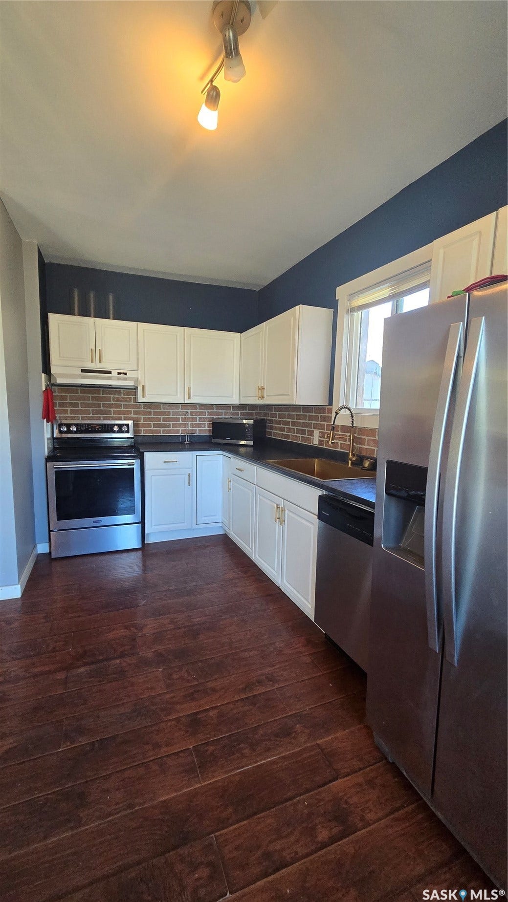 White cabinetry with gold-tone hardware, stainless steel appliances, brick-patterned tile backsplash, dark countertops, and wood-finish flooring