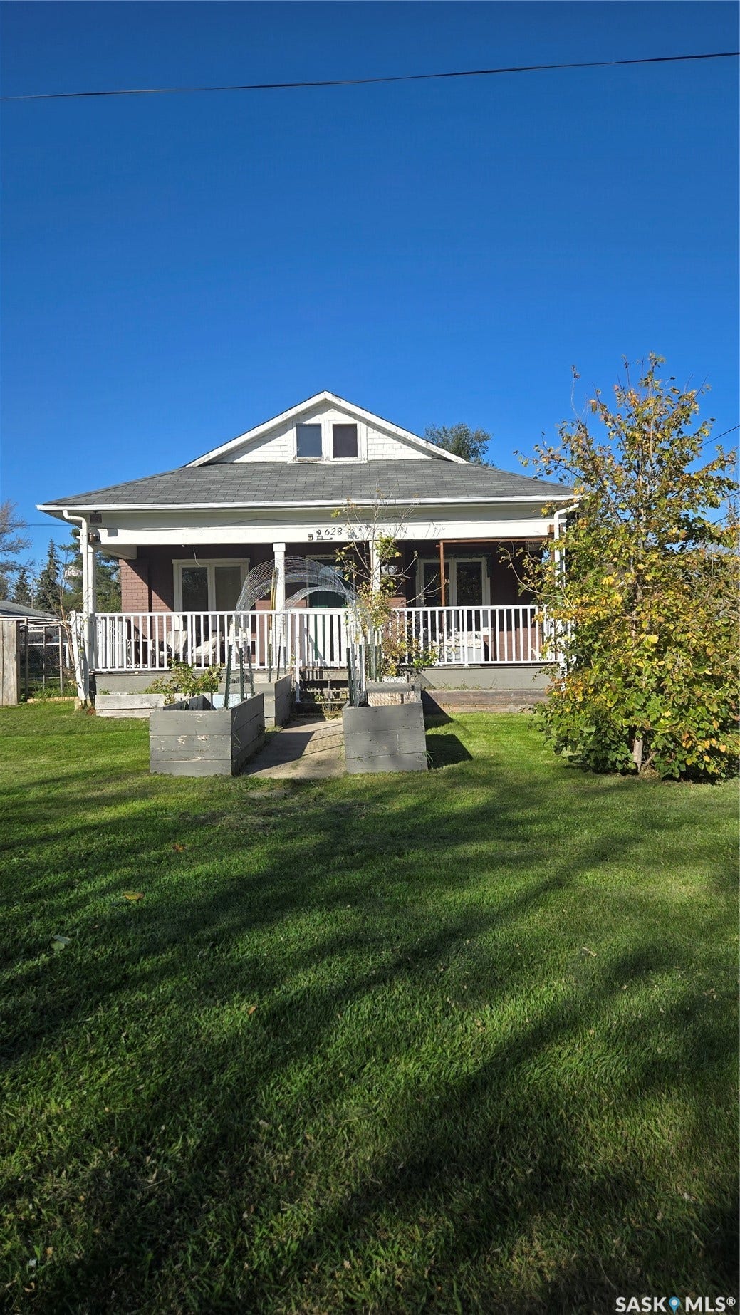 Gabled roofline with dormer windows, full-width covered porch, white railing, and raised garden beds