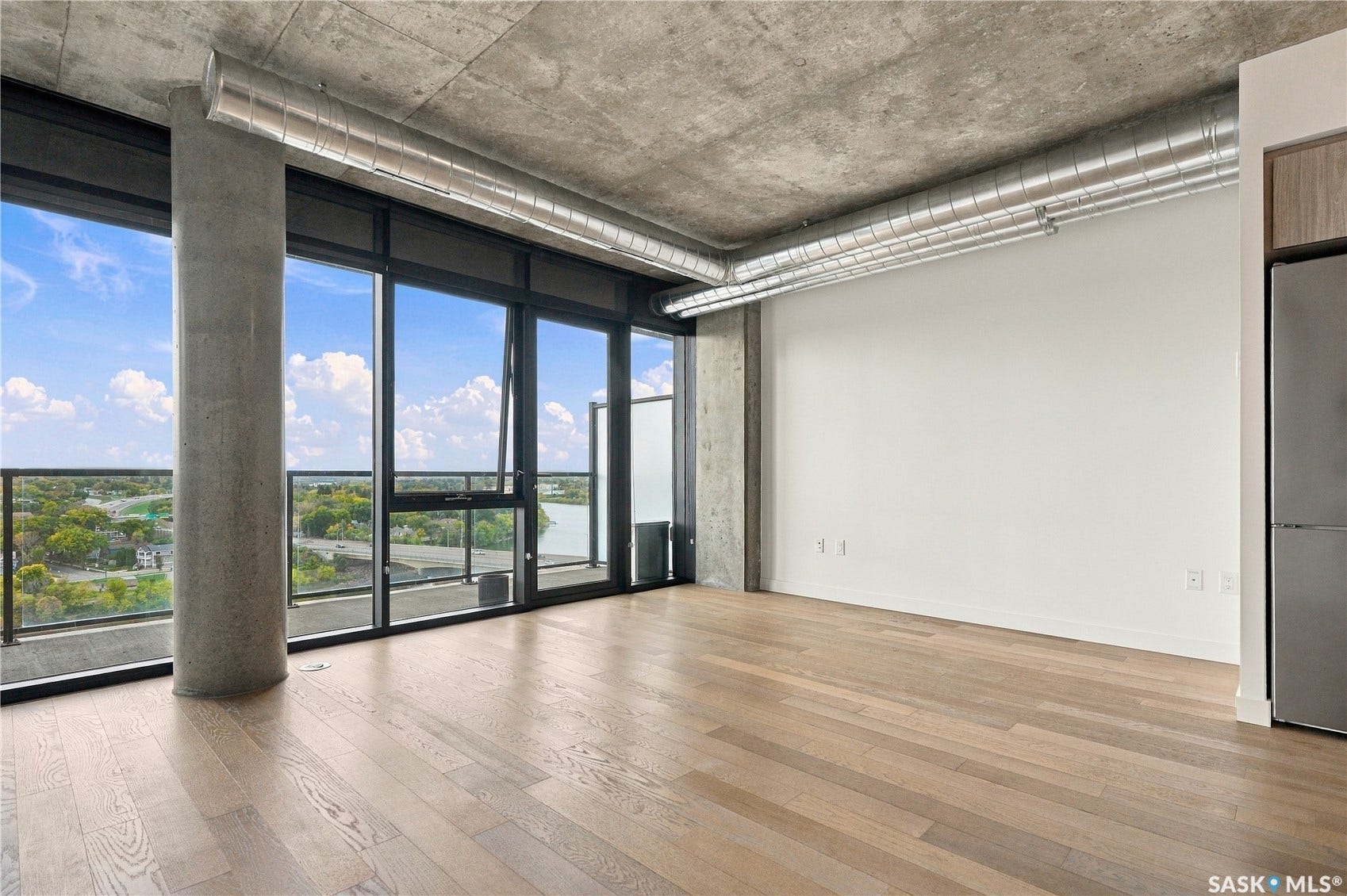Floor-to-ceiling windows, light wood-finish flooring, exposed concrete ceiling, exposed ductwork, and a partial built-in stainless steel refrigerator