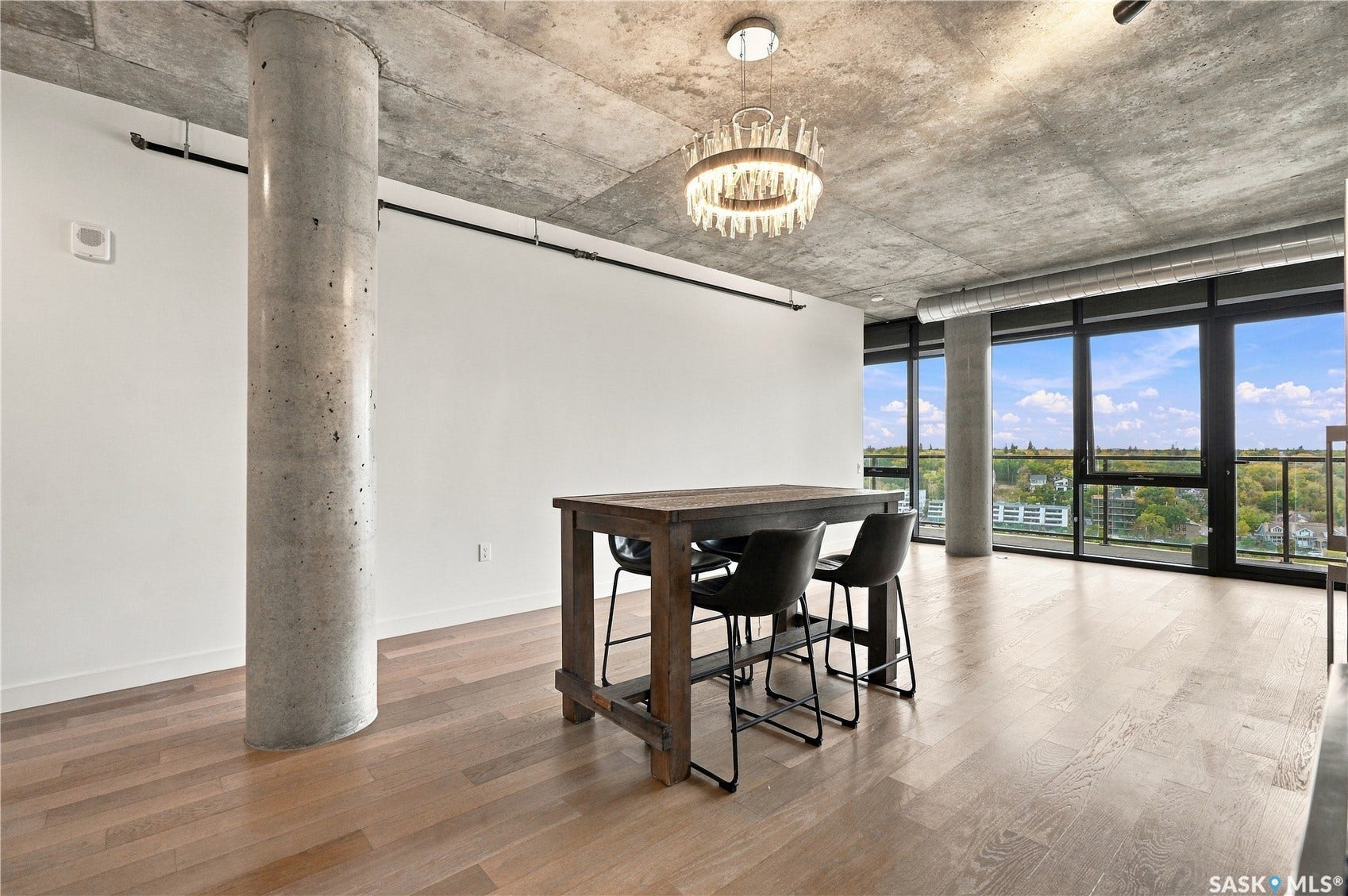 Expansive floor-to-ceiling windows, exposed concrete ceiling, light wood-finish flooring, and a contemporary chandelier