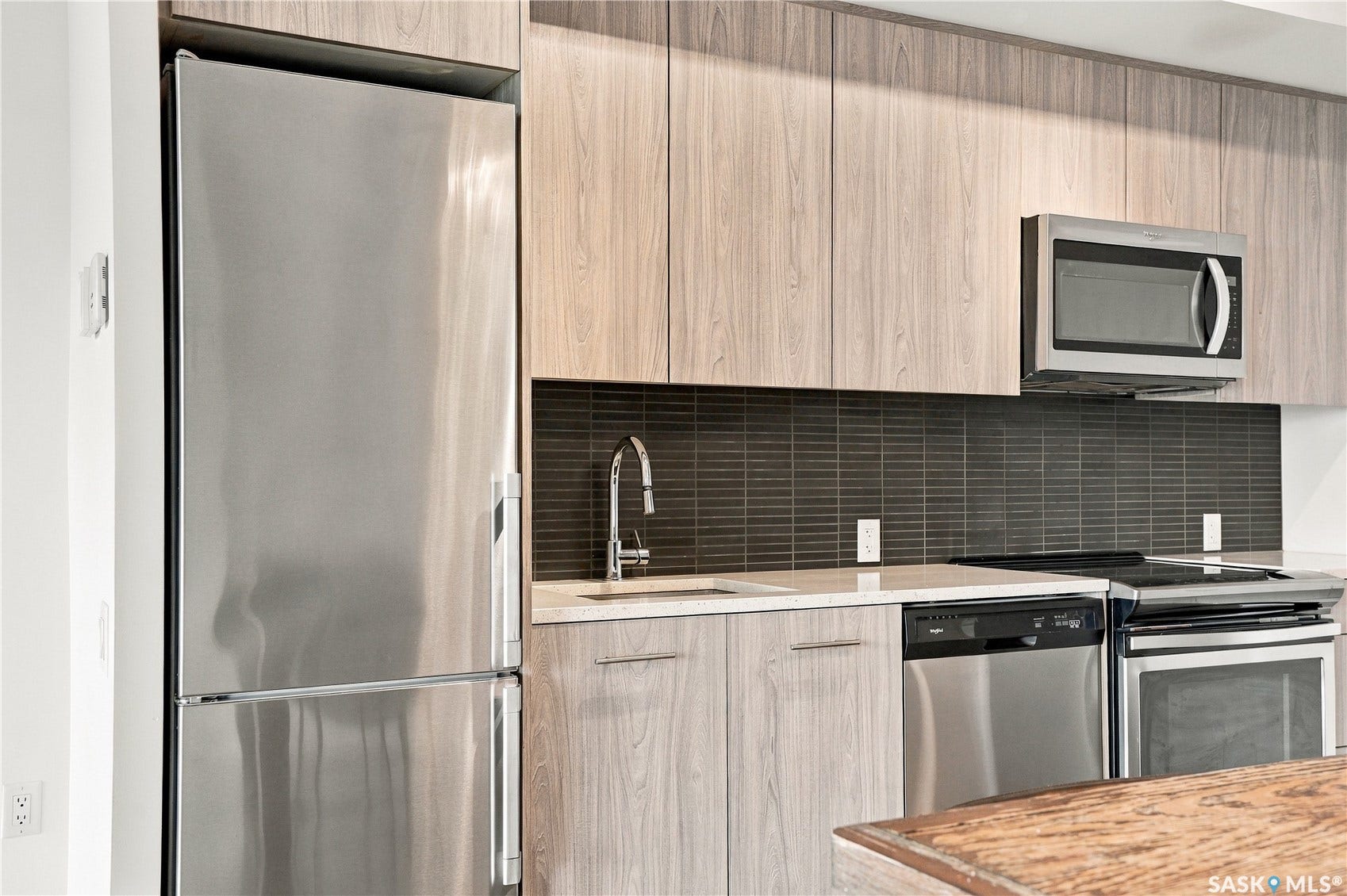 Stainless steel appliance suite, sleek wood-finish cabinetry, dark tile backsplash, and light countertops