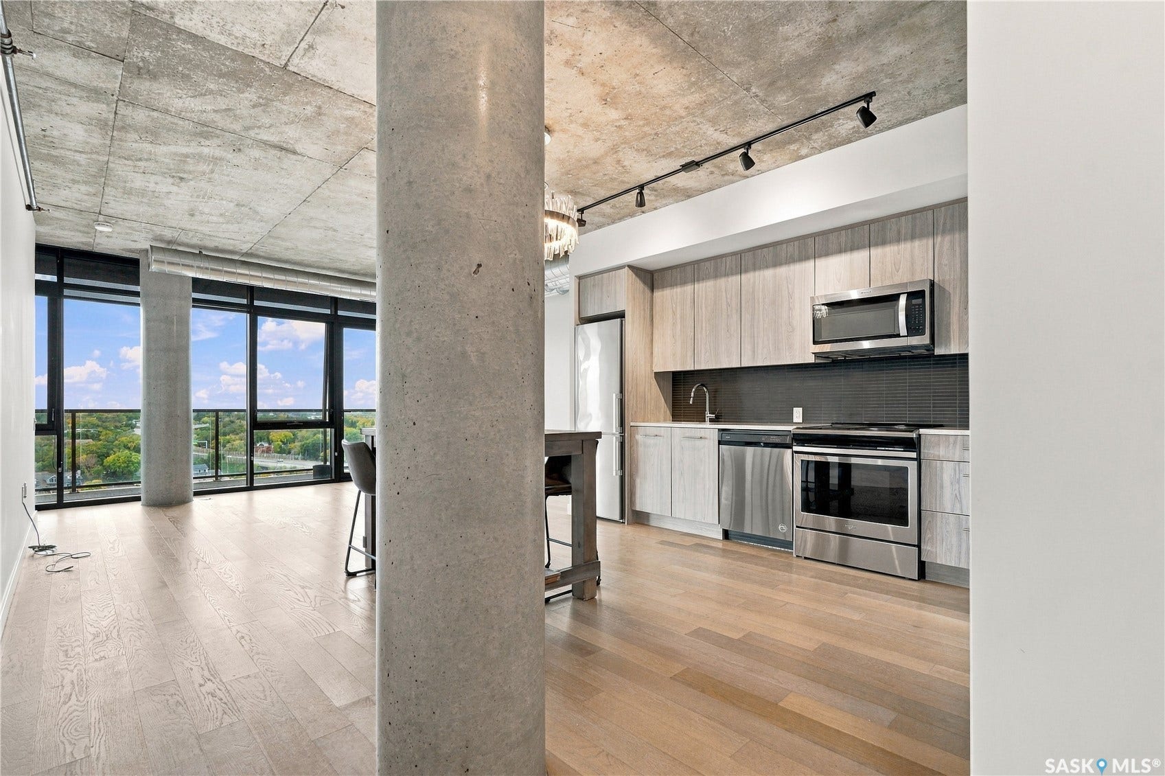 Floor-to-ceiling windows, exposed concrete ceiling, light wood-finish flooring, and a kitchen featuring stainless steel appliances and wood-finish cabinetry