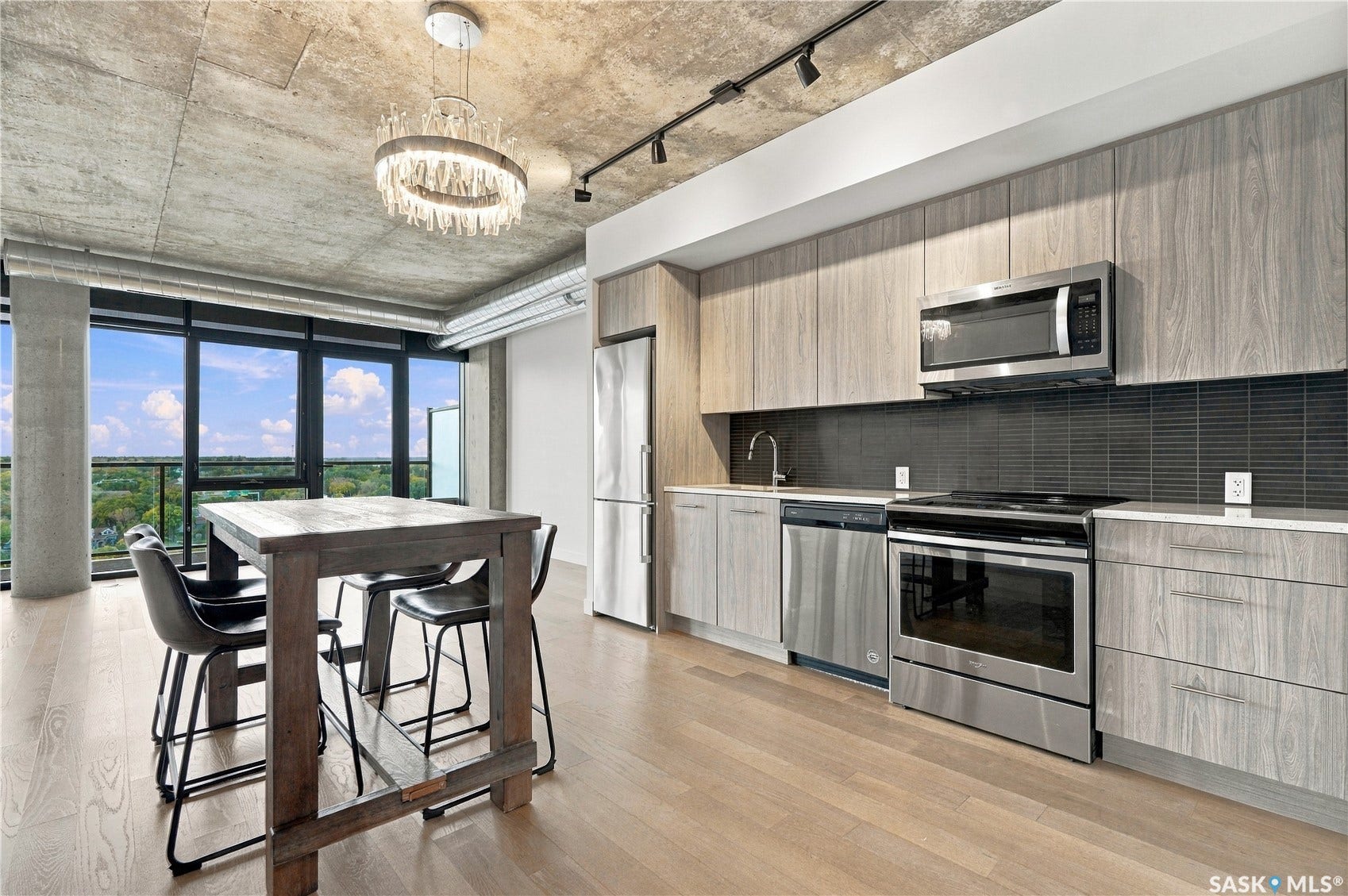 Concrete ceiling with track lighting and a decorative chandelier, expansive windows, light wood-finish flooring, and a kitchen featuring stainless steel appliances with wood-finish cabinetry