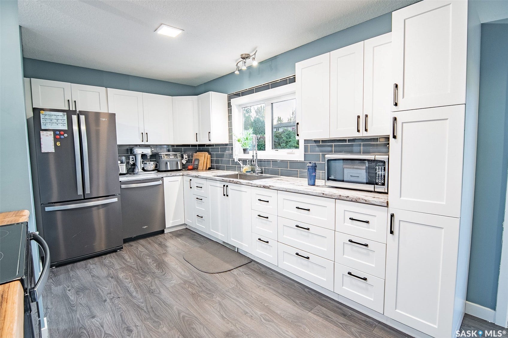 White cabinetry with matte black hardware, grey subway tile backsplash, wood-finish flooring, stainless steel appliances, and a window above the sink