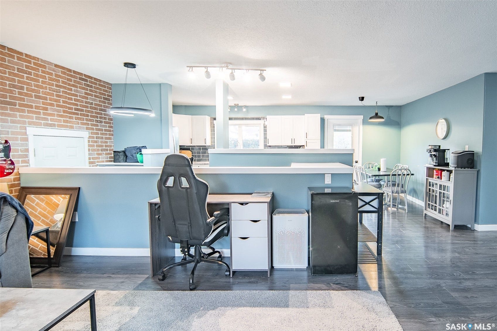 Open-concept layout with brick accent wall, wood-finish flooring, white cabinetry, and track lighting