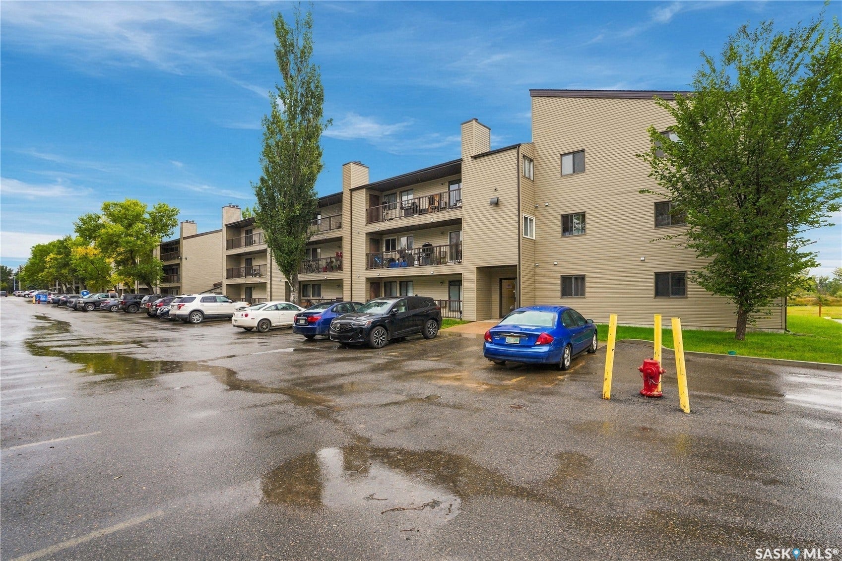 Multi-story building featuring multiple private balconies, a paved parking area, and surrounding mature trees