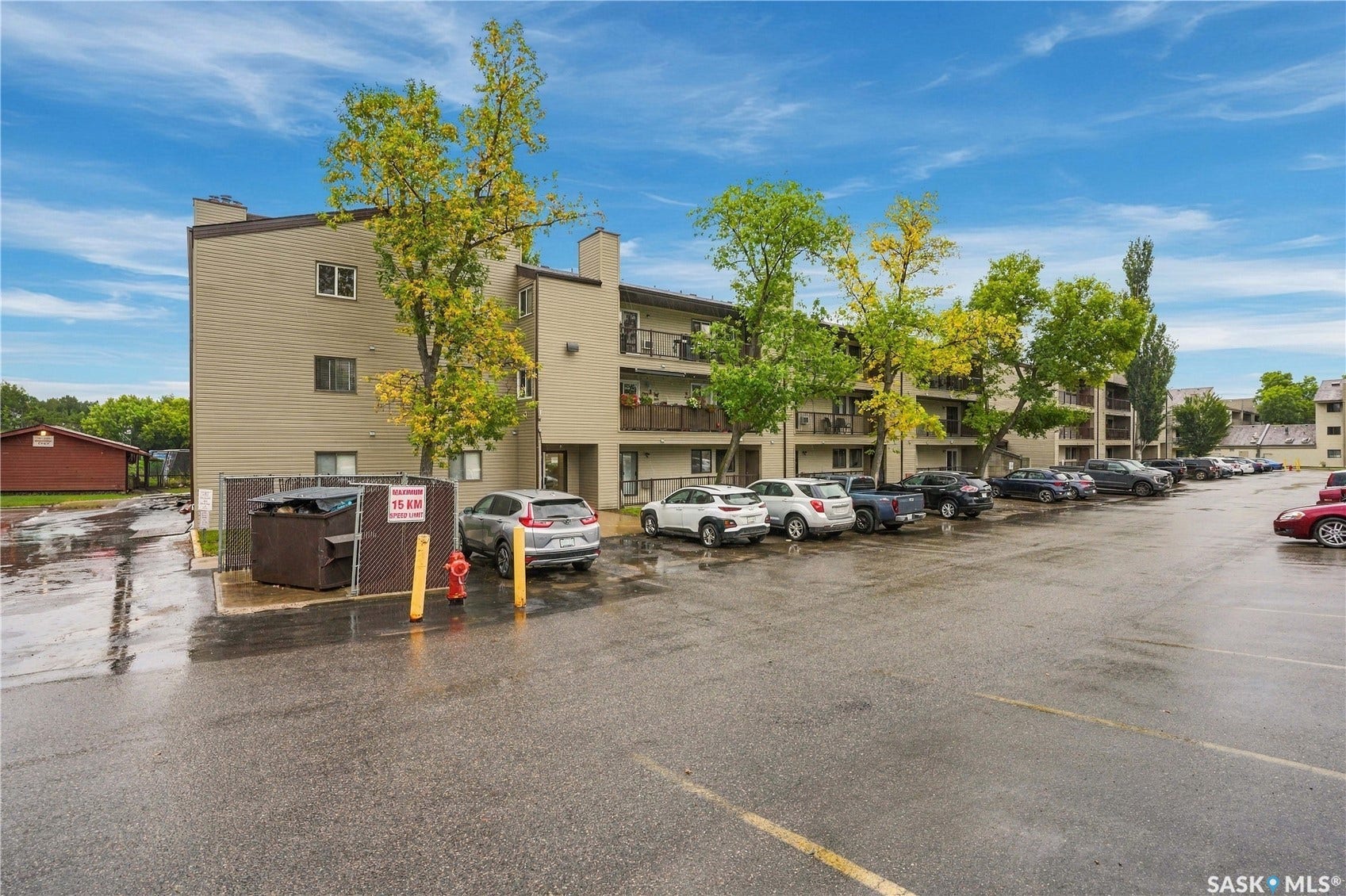 Multi-story building with balconies, surface parking area, and mature trees
