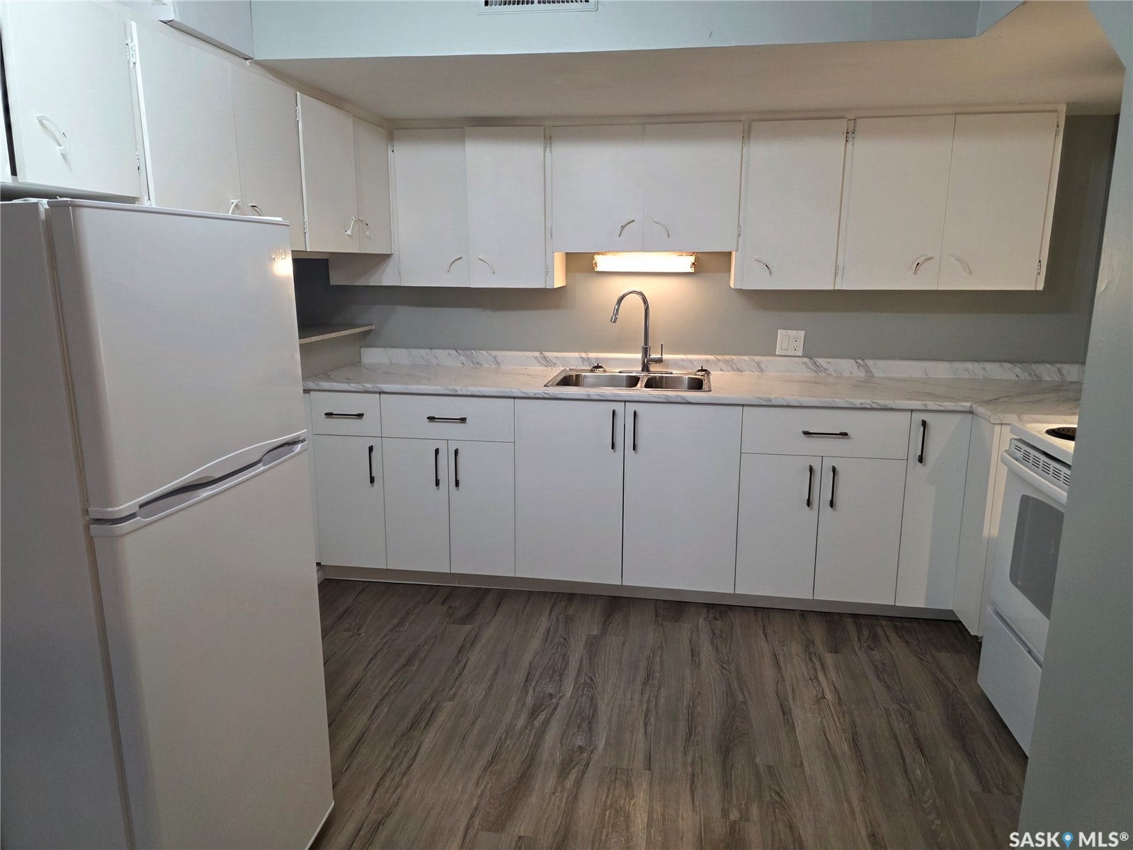 White cabinetry with black hardware, wood-finish flooring, undermount sink with gooseneck faucet, light-toned countertops, and included refrigerator