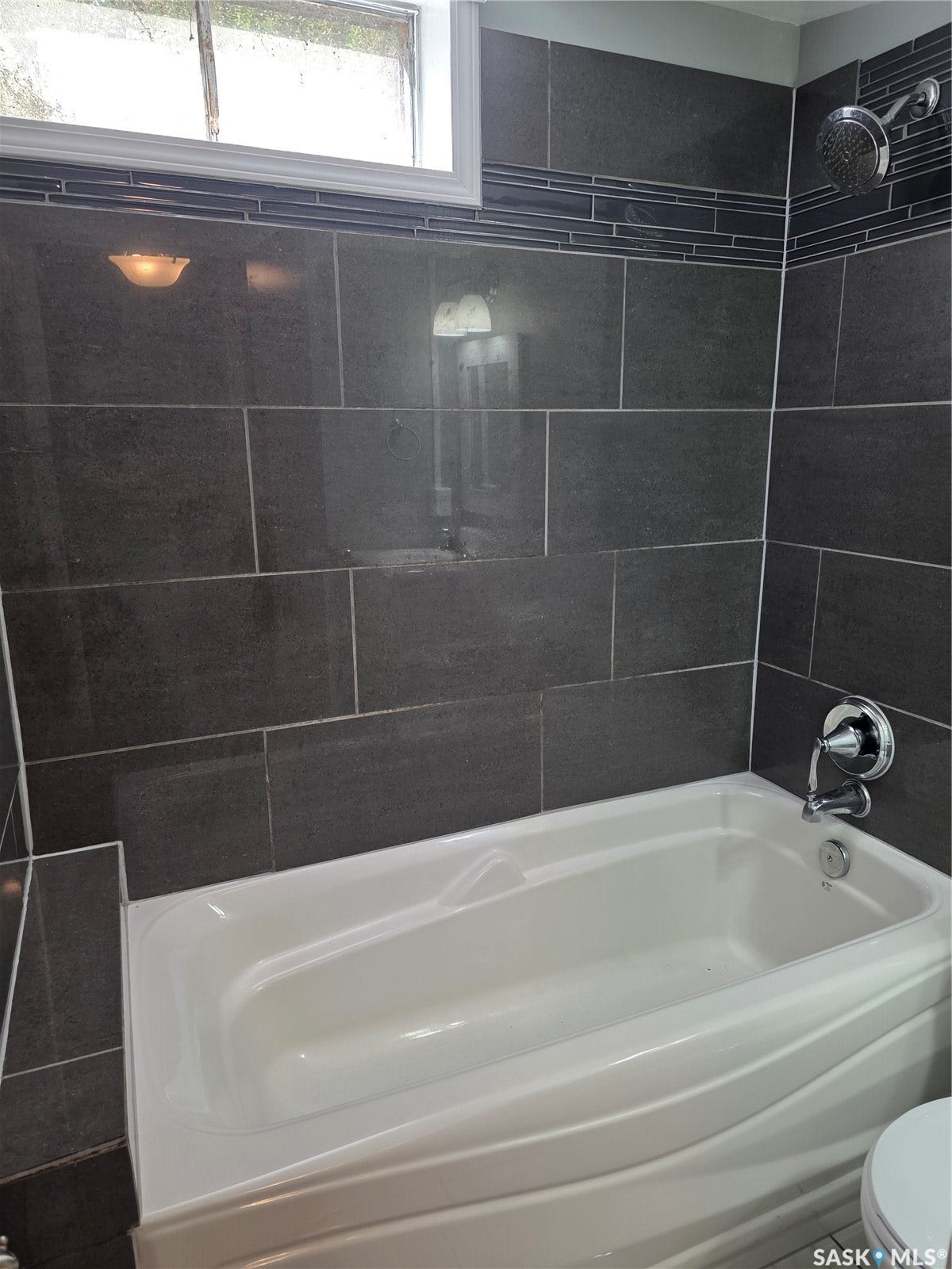 Soaking tub with chrome fixtures, surrounded by large format dark gray tile, accented with a decorative tile band, and a window for natural light