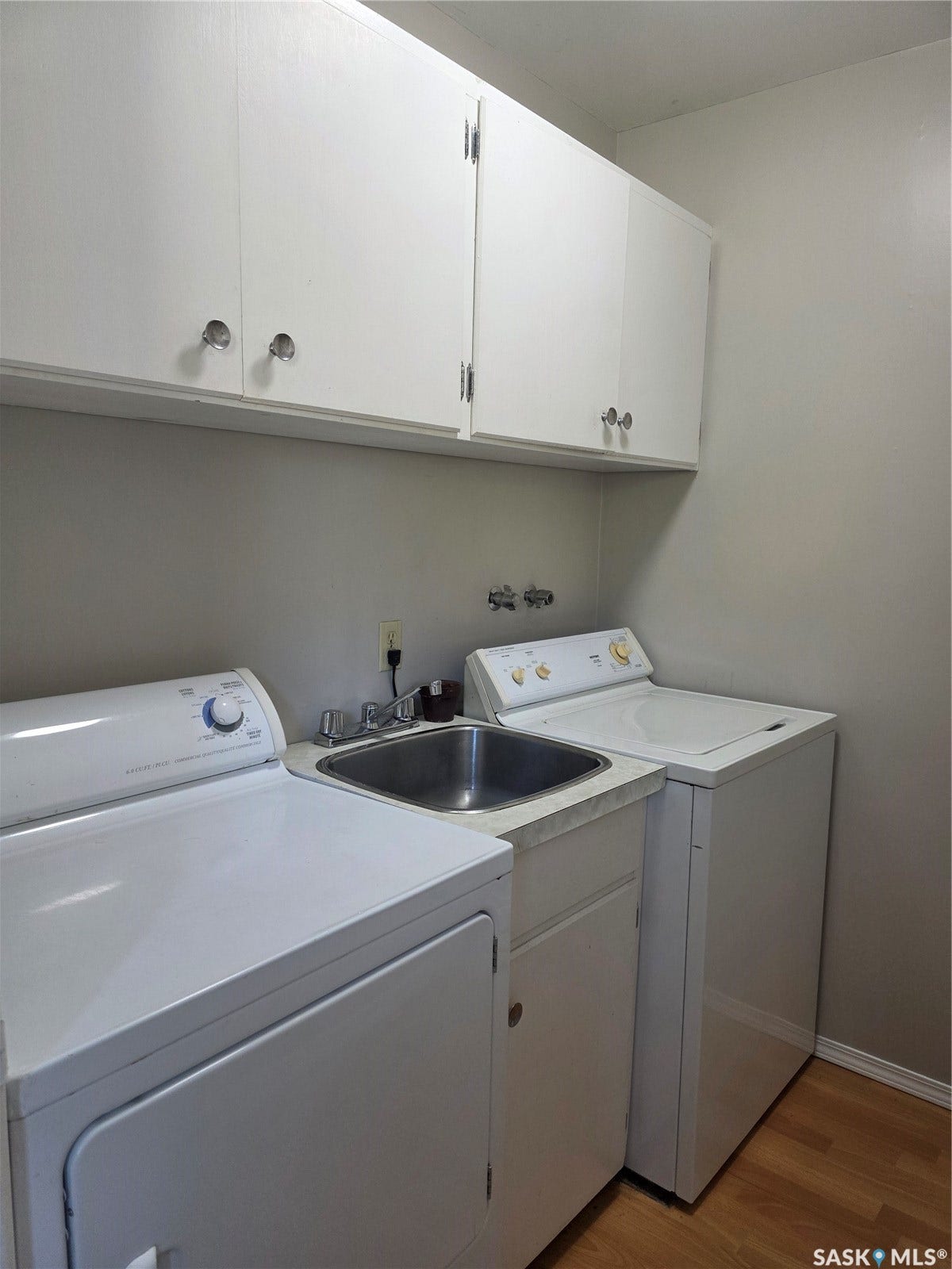 Wood-finish flooring with white wall-mounted cabinetry, a stainless steel utility sink, and a built-in counter