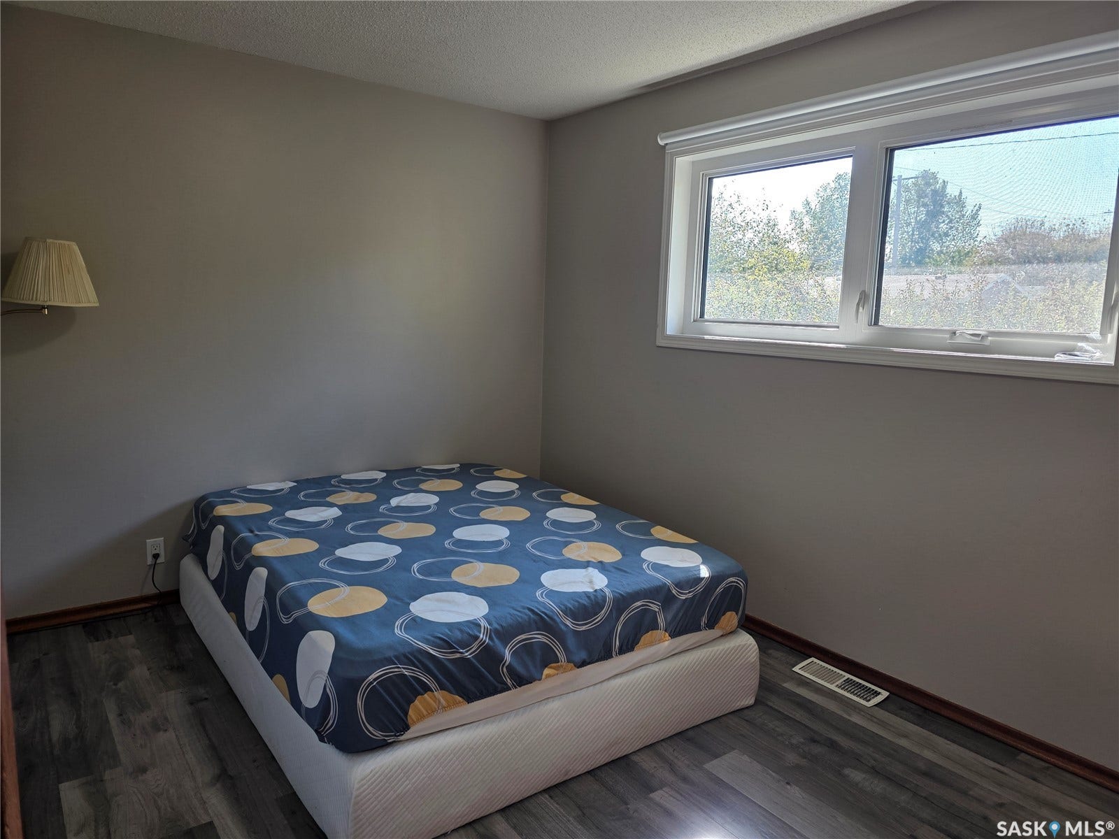 Wood-finish flooring with baseboard trim, wall-mounted light fixture, and a window with roller shade