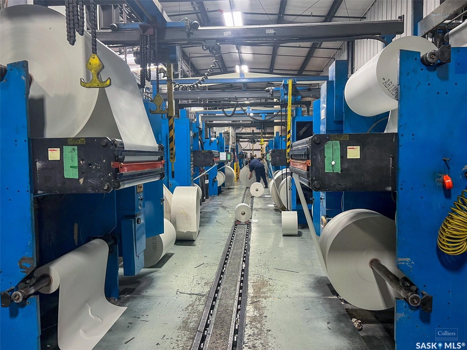 Industrial interior with multiple large-scale blue machinery units, overhead gantry system, and wide roll material feeding into equipment