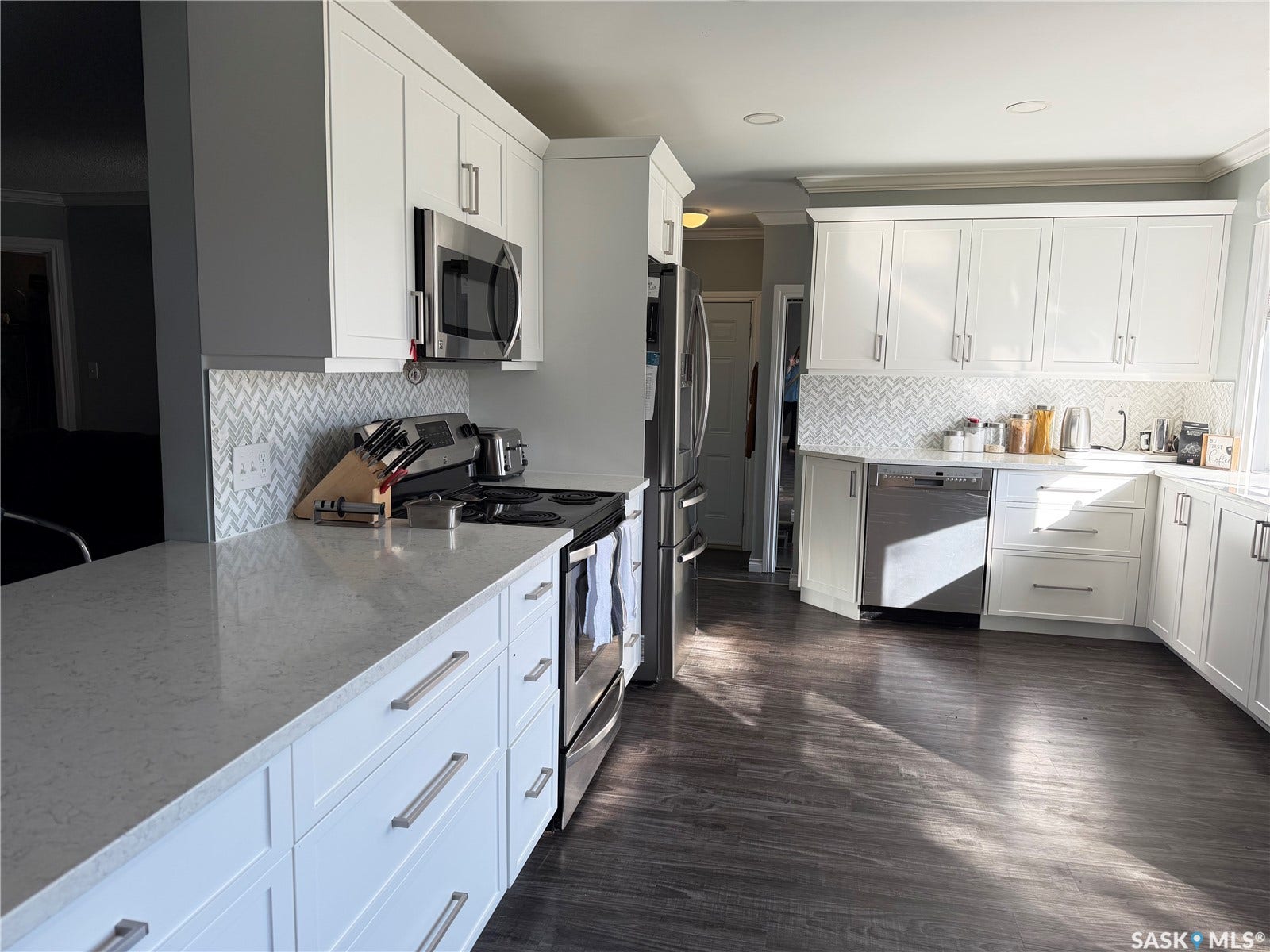 White cabinetry with stainless steel hardware, light-toned countertops, herringbone pattern tile backsplash, and wood-finish flooring