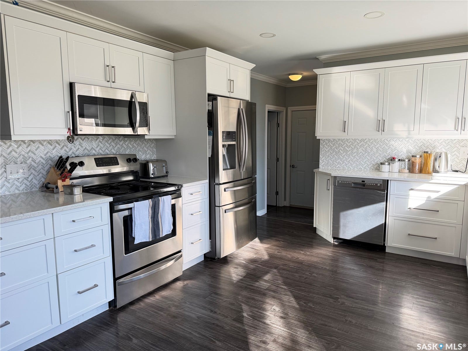 White shaker-style cabinetry, stainless steel appliances, light-toned countertops, herringbone pattern tile backsplash, and dark wood-finish flooring