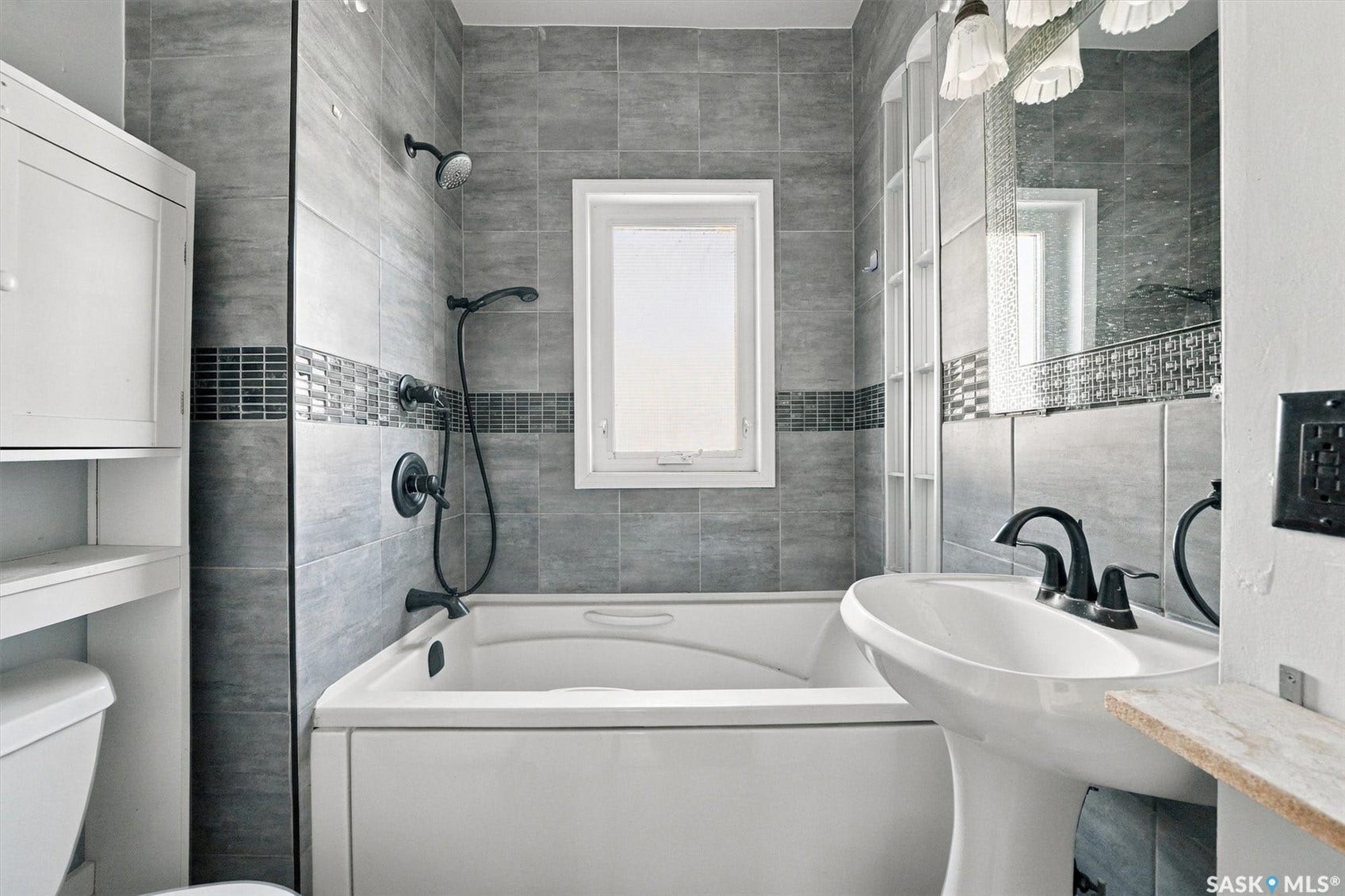 Bathtub with matte black fixtures, grey tiled walls with mosaic accent, pedestal sink, and built-in storage cabinet